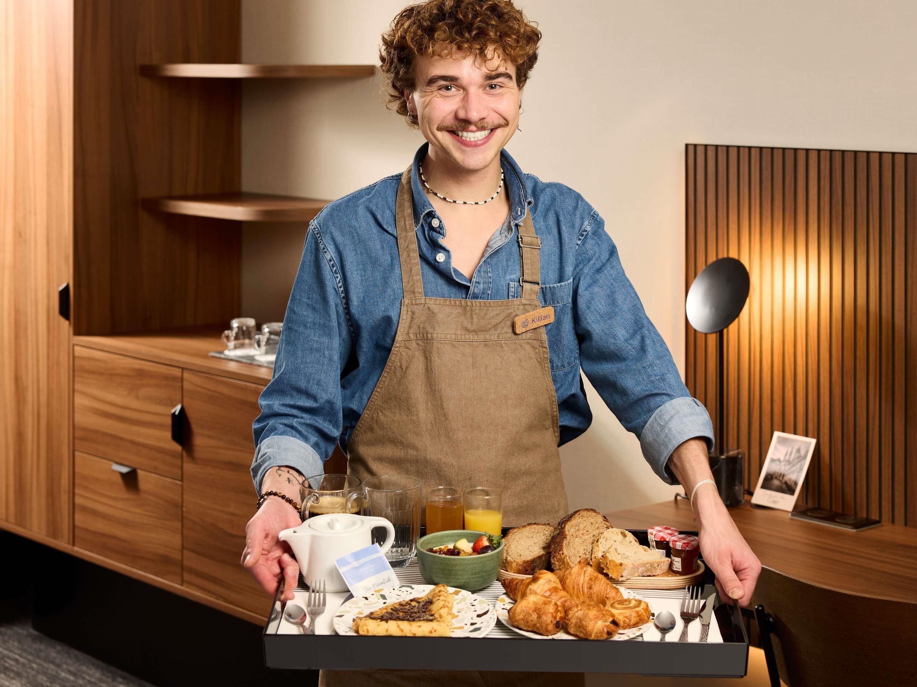 Uma pessoa servindo café da manhã com uma bandeja contendo croissants, pão, uma tigela de frutas e um bule.