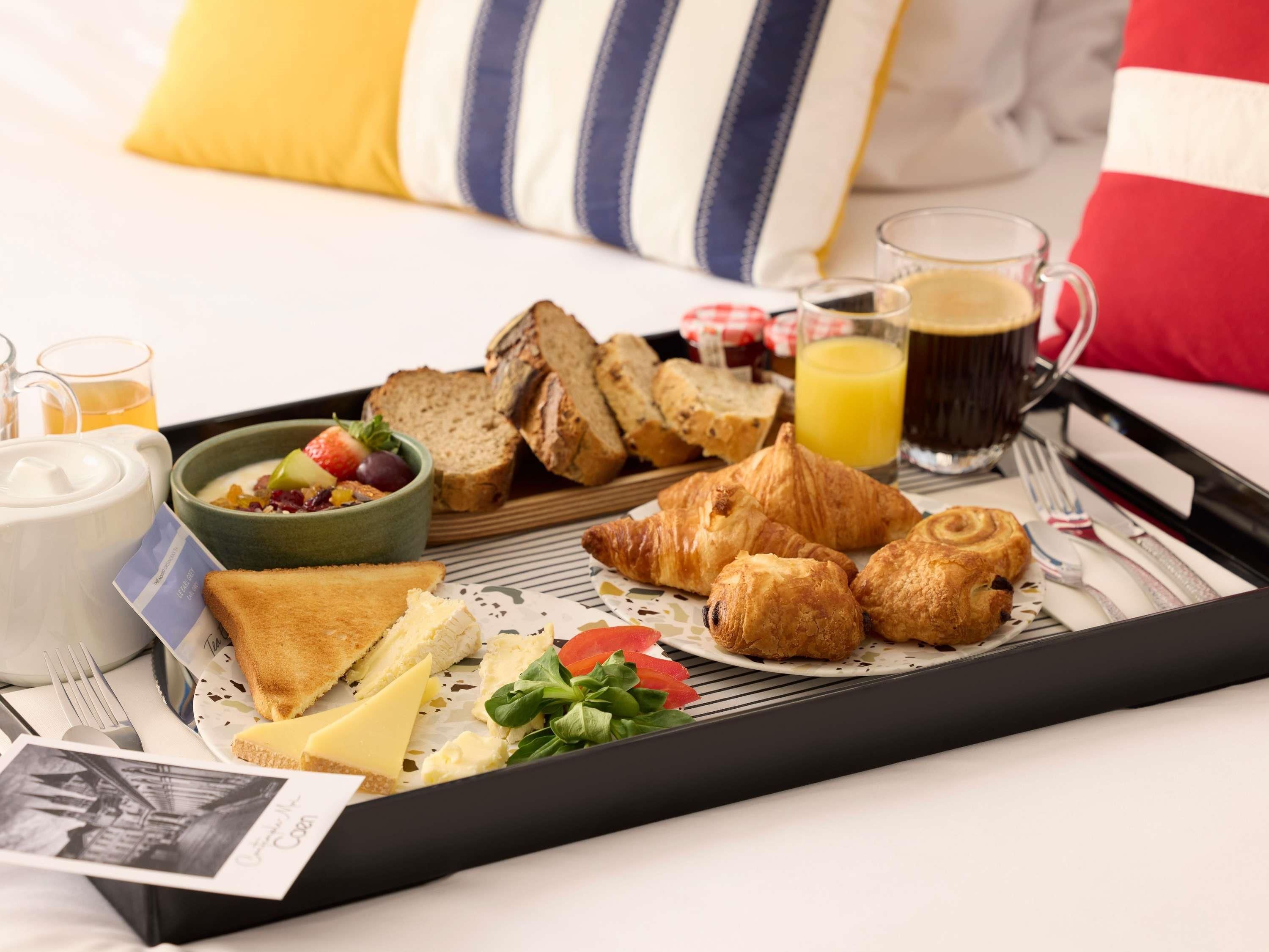 A breakfast tray with croissants, bread, cheese, and a bowl of fruit on a bed with pillows.