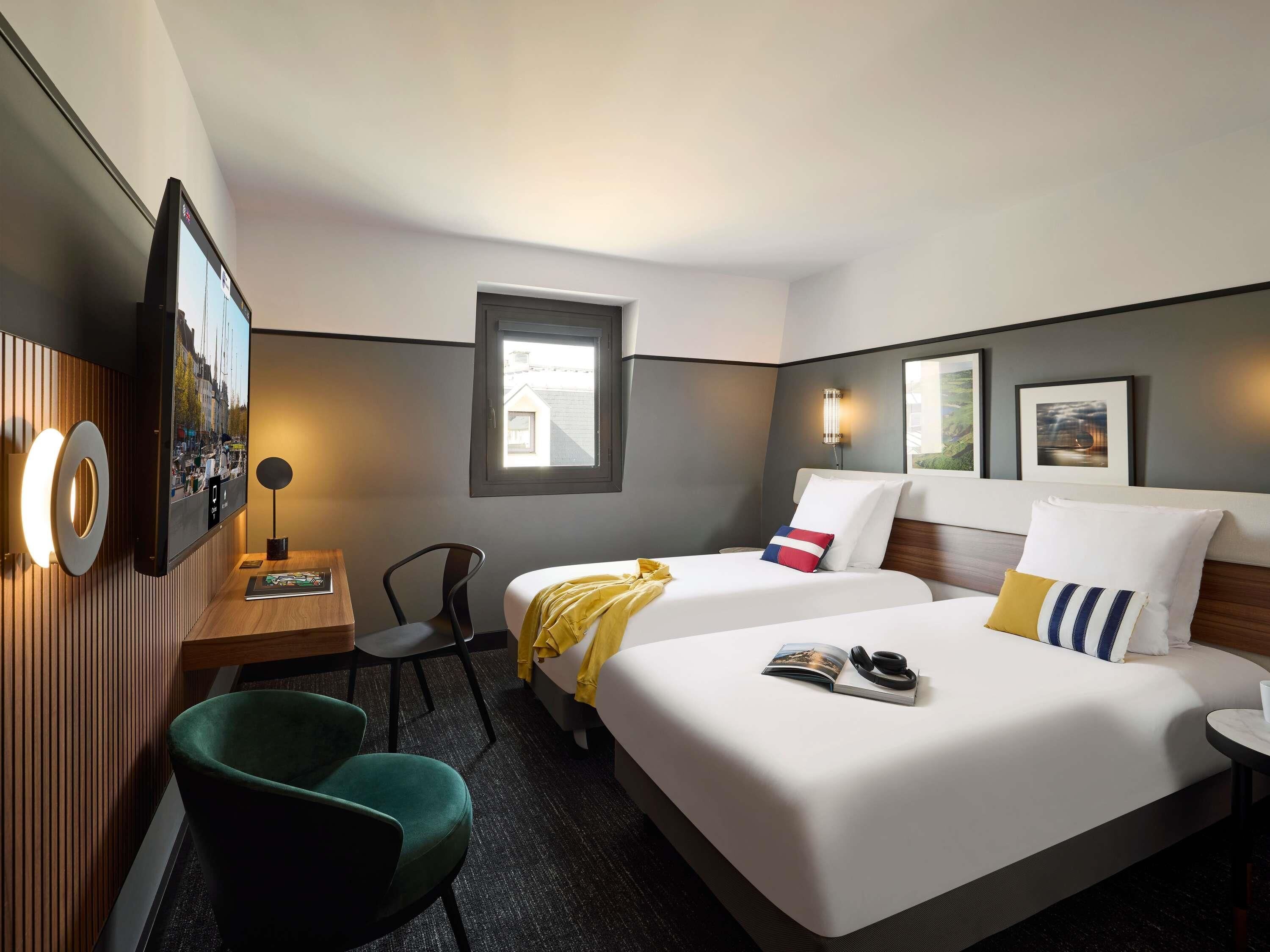 A hotel room with a bed, a desk, a chair, a window, and wall-mounted artwork.
