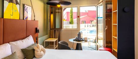 Habitación de hotel con cama, una mesita con hervidor, una silla y vistas al exterior.
