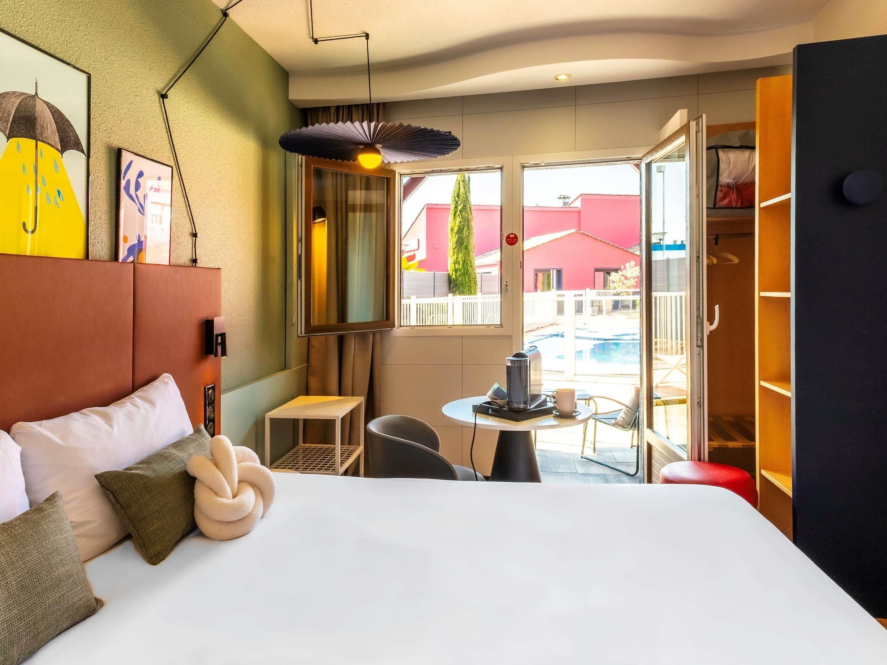 A hotel room with a bed, a small table with a coffee maker, a chair, and a view of a swimming pool and buildings.