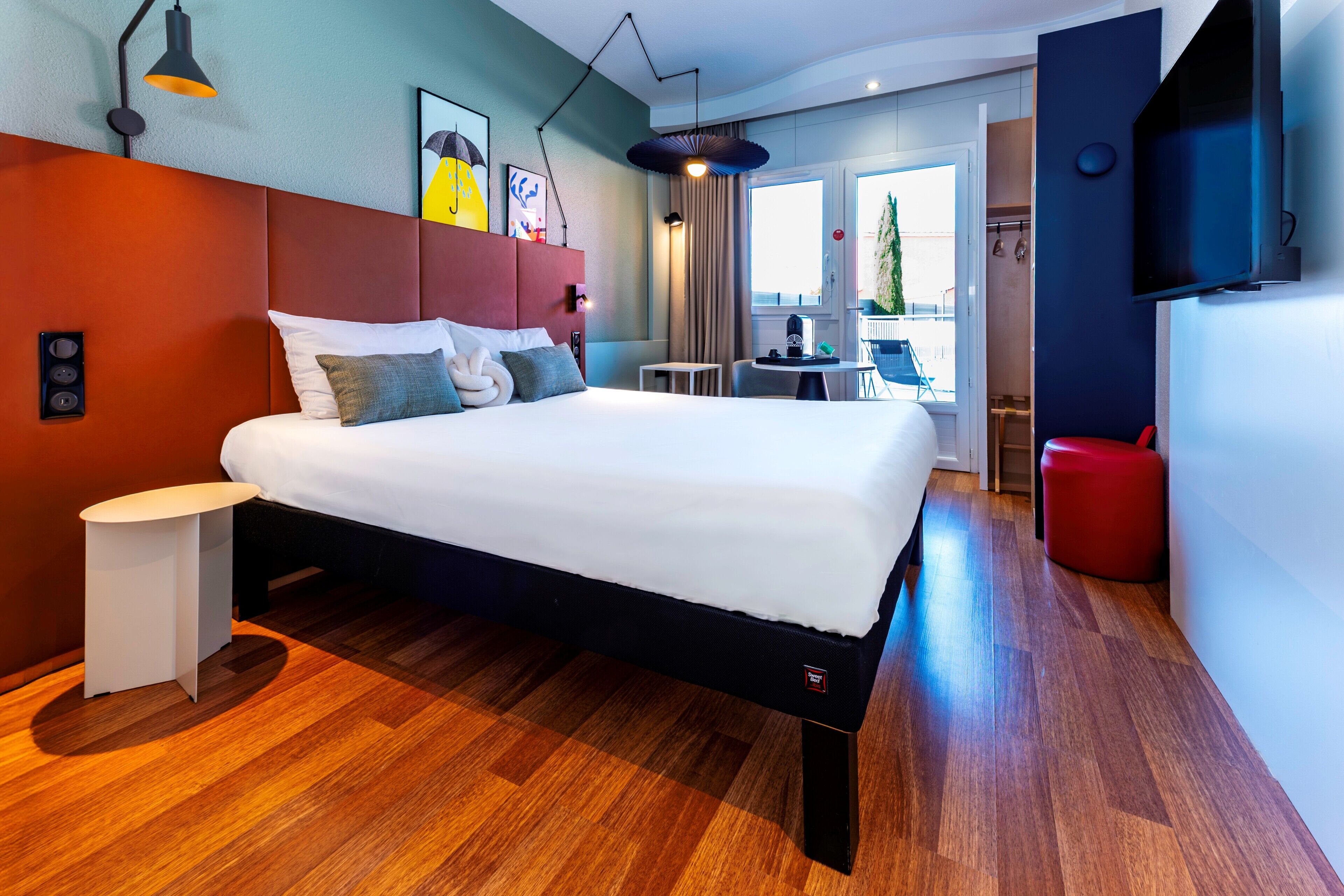 A modern hotel room with a large bed, a small table, a red chair, and a TV mounted on the wall.