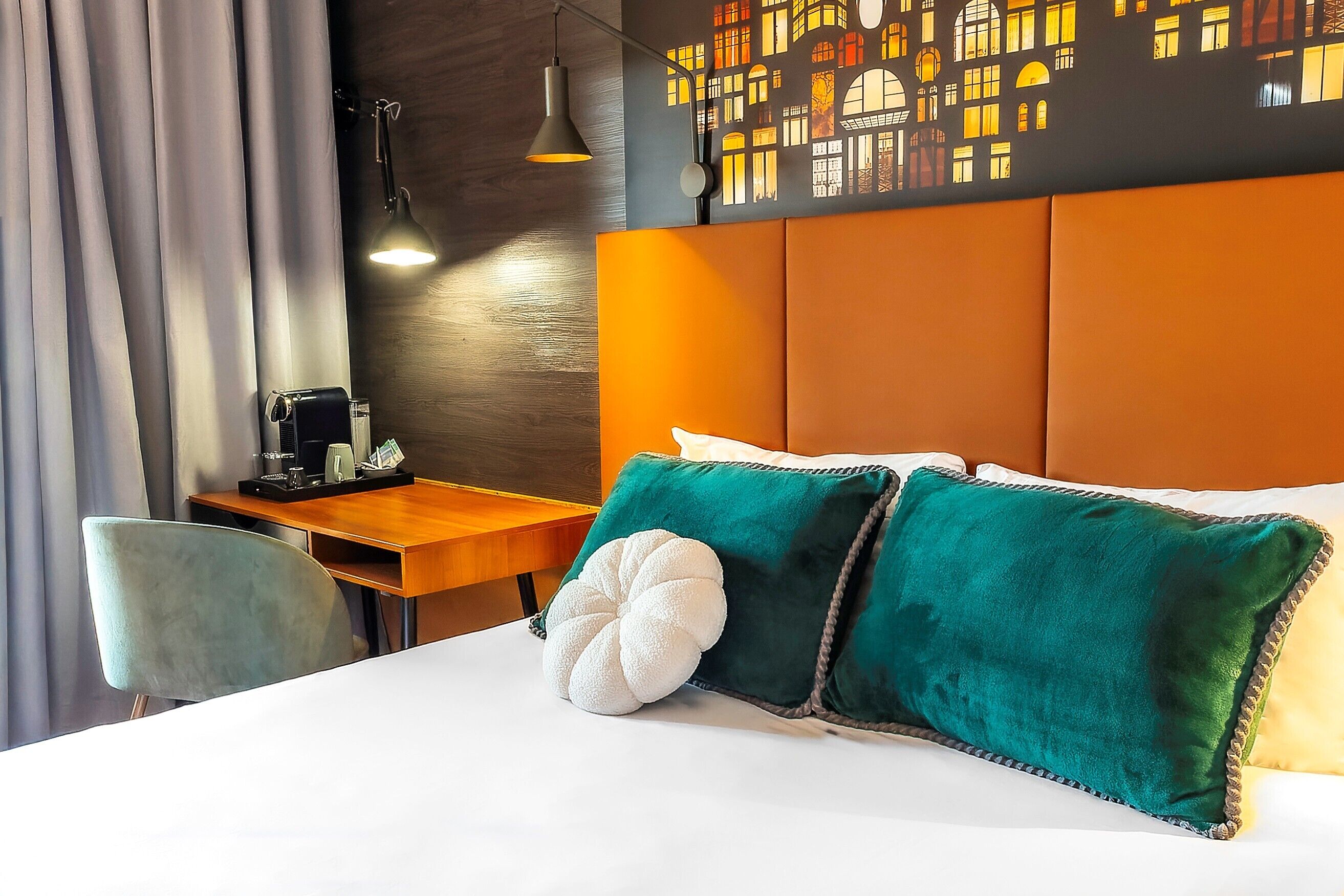A hotel room with a bed, a desk with a coffee maker, a chair, and a wall with a cityscape mural.