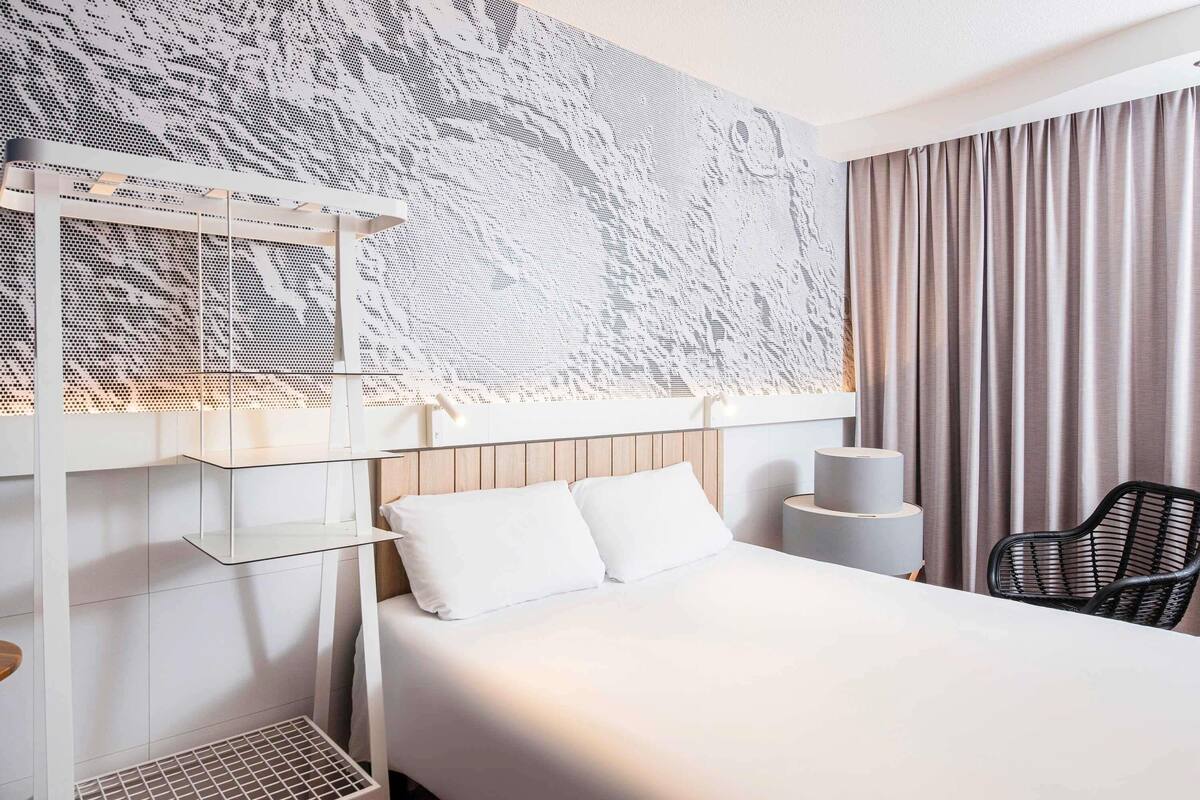 A hotel room with a bed, a chair, a small table, and a wall-mounted shelf.