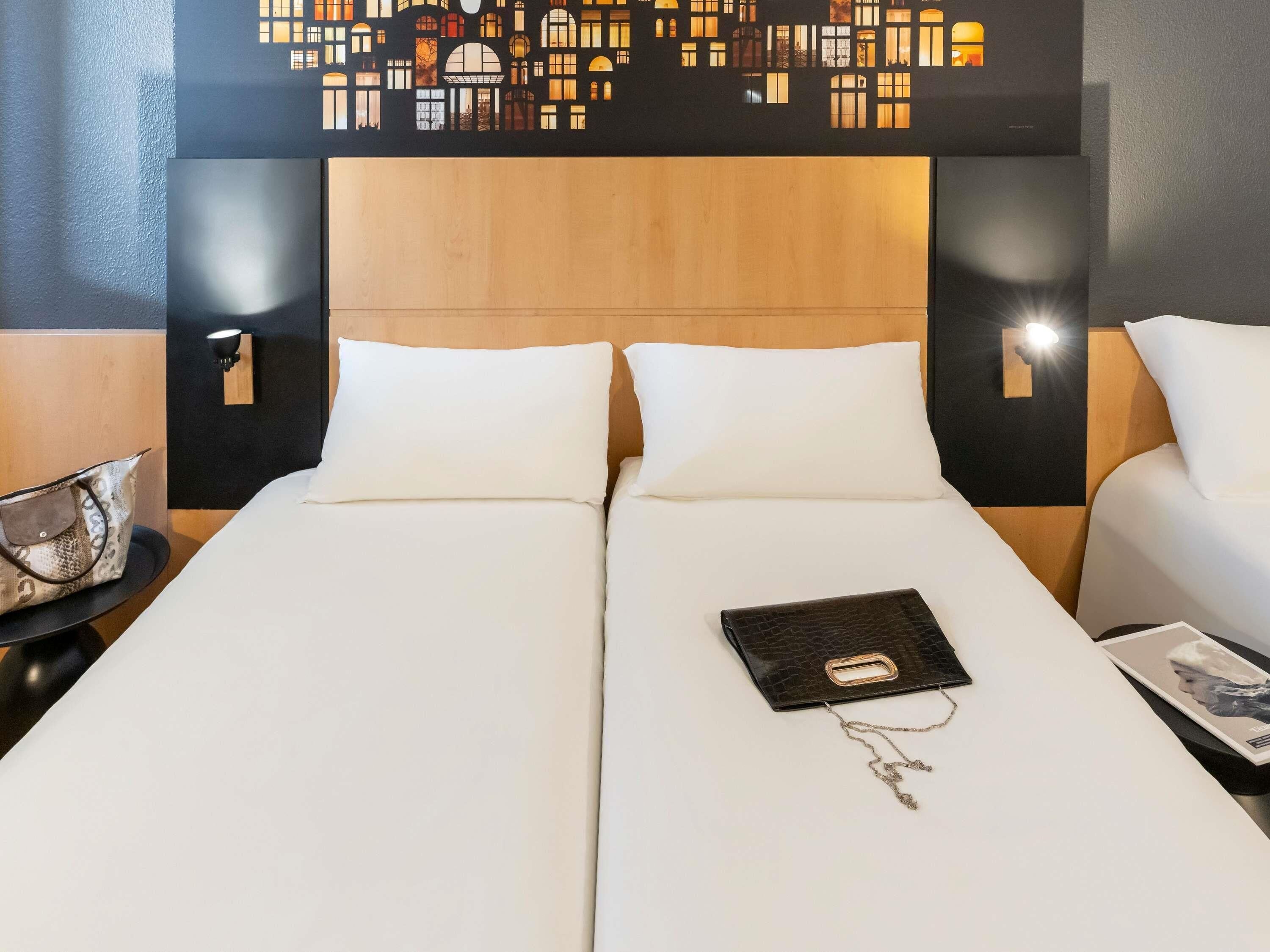 Una cama doble con sábanas blancas, un bolso de cuero negro sobre la cama y un panel decorativo en la pared.