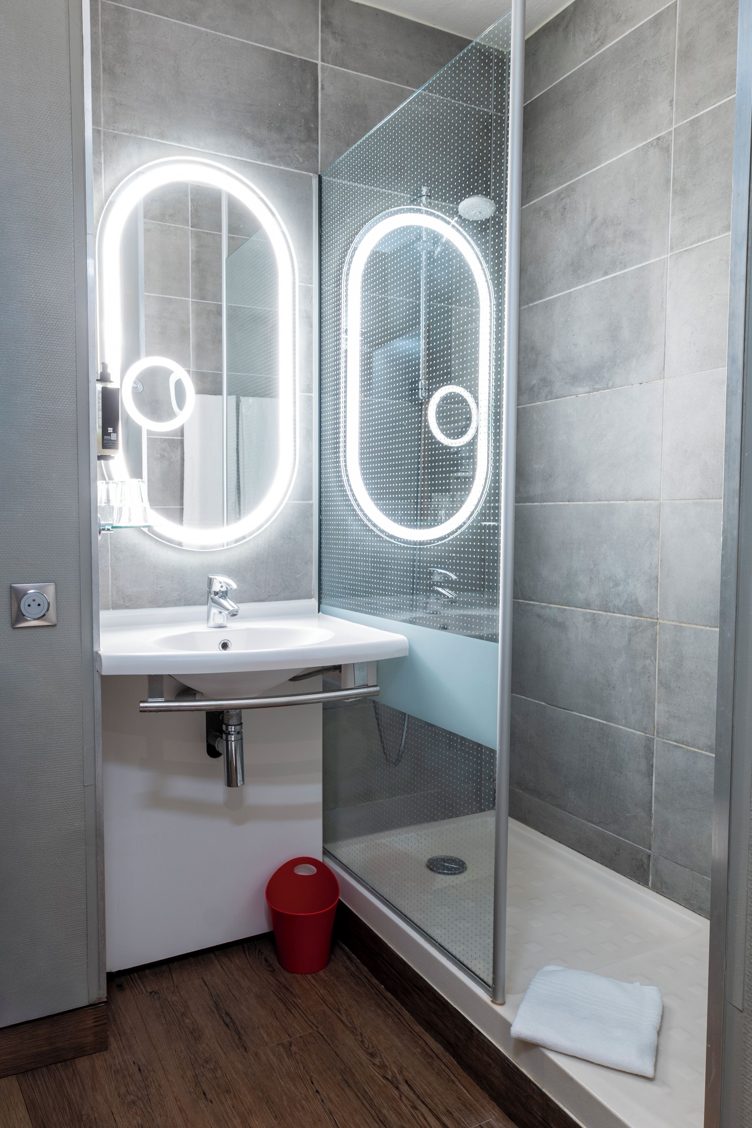 A modern bathroom with a glass-enclosed shower, a white sink with a mirror, and a red trash bin.