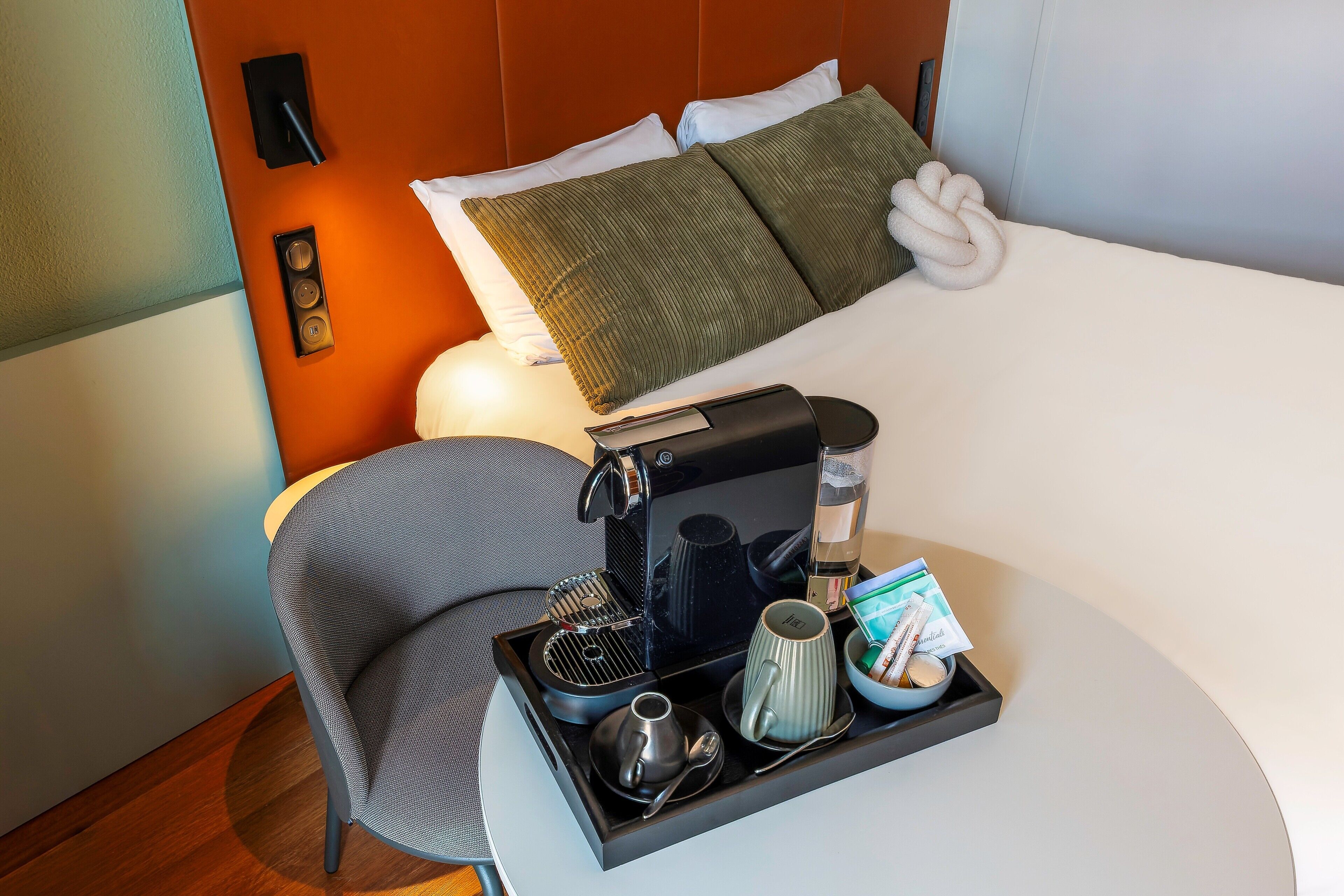 Habitación de hotel con una cama, una mesita de noche con cafetera, una bandeja con taza y un bol, y una silla.