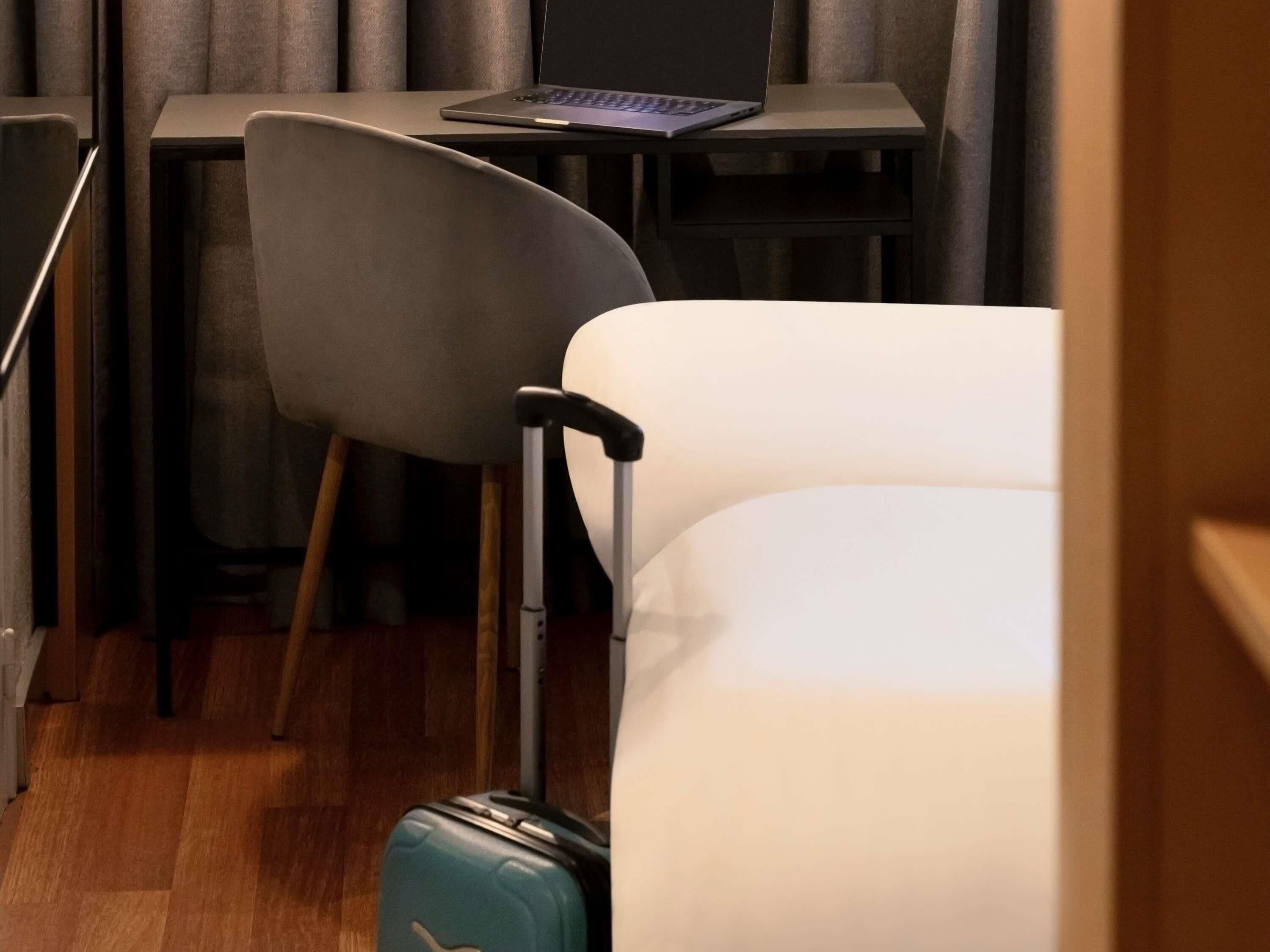 A hotel room with a bed, a desk with a laptop, a chair, and a suitcase.