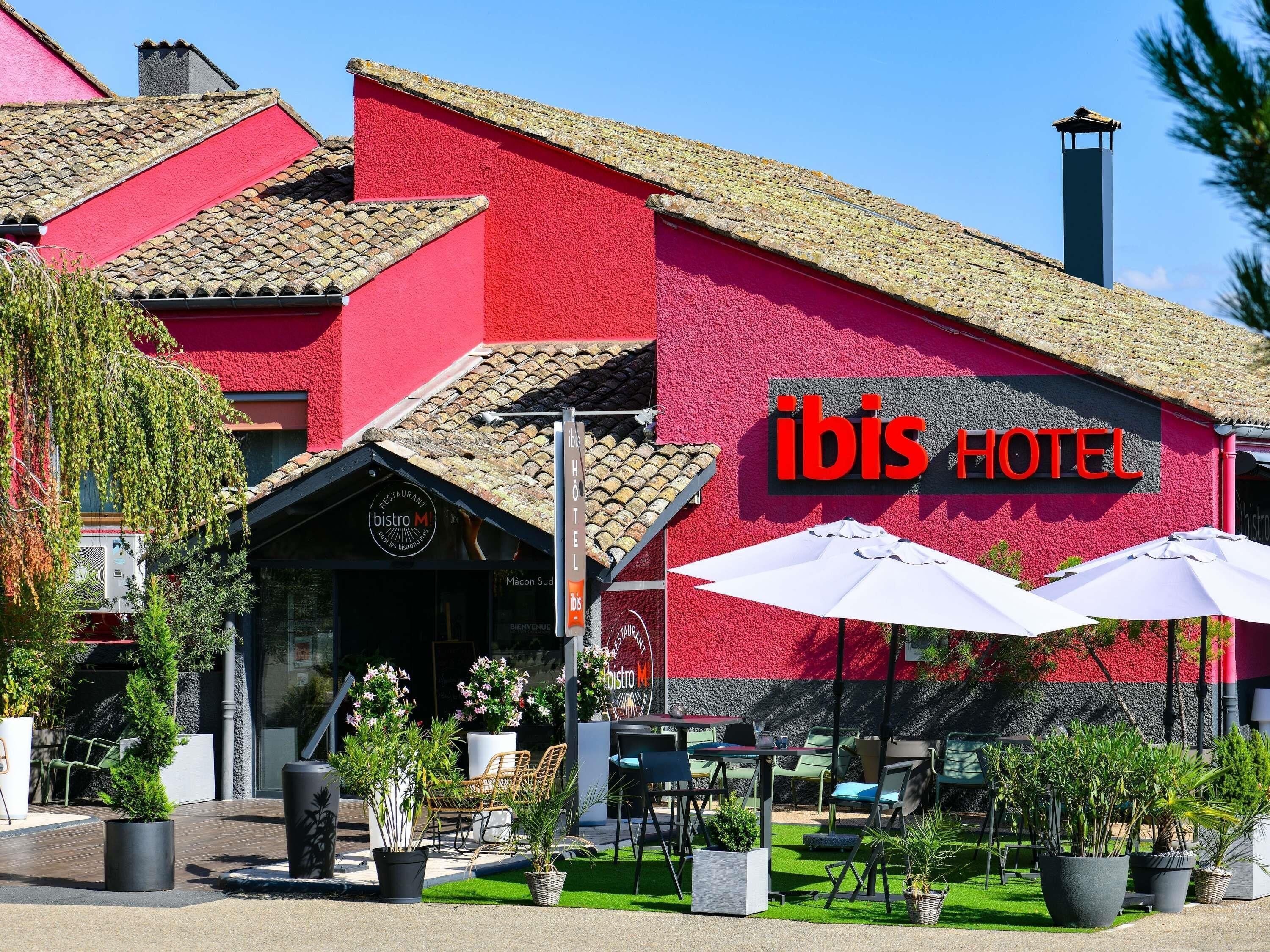 Un edificio rojo con un letrero que dice 'ibis HOTEL' y asientos exteriores con sombrillas.