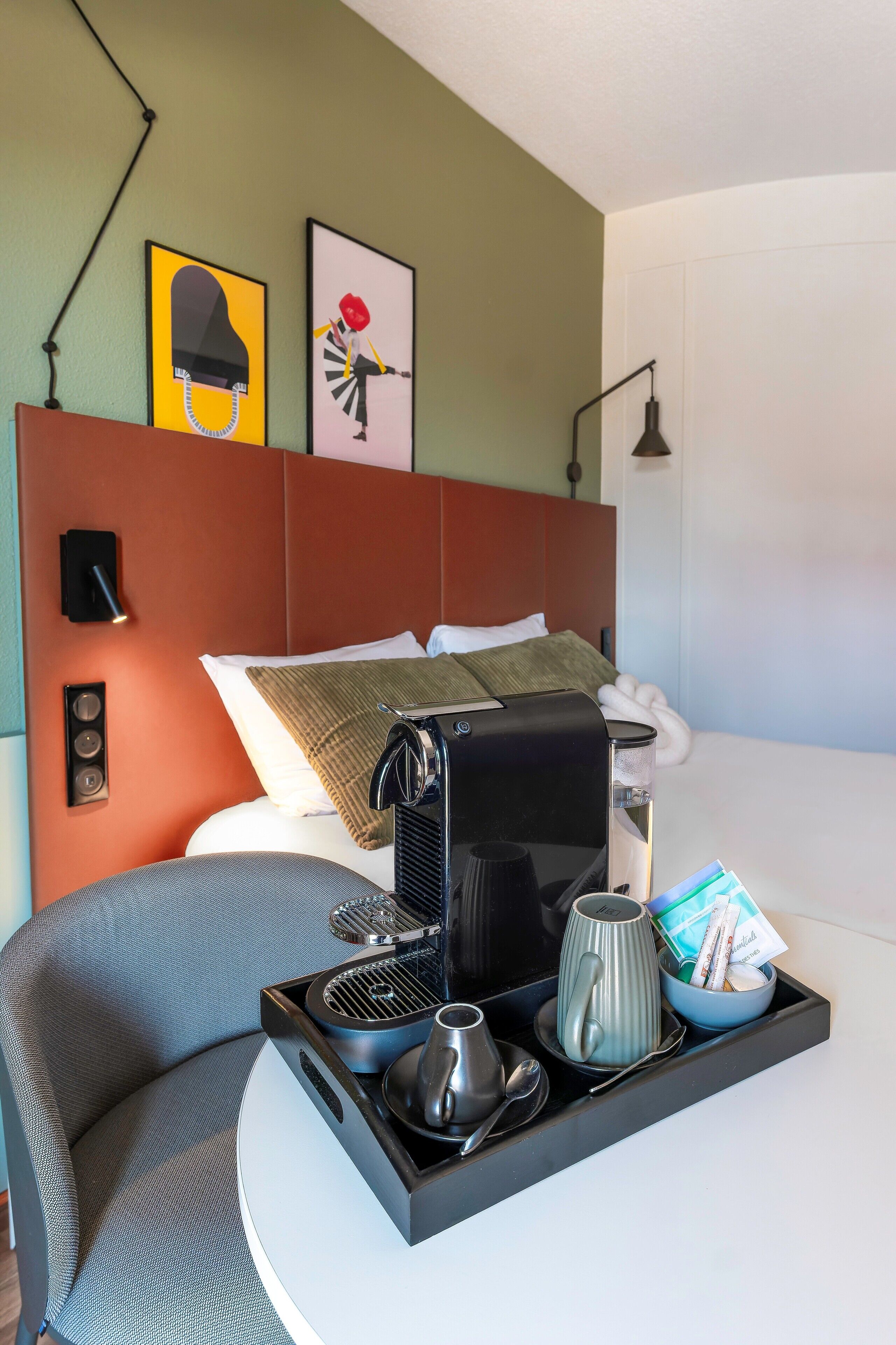Habitación de hotel con cama, cafetera, bandeja con tazas y una taza, almohada y dos lámparas de pared.