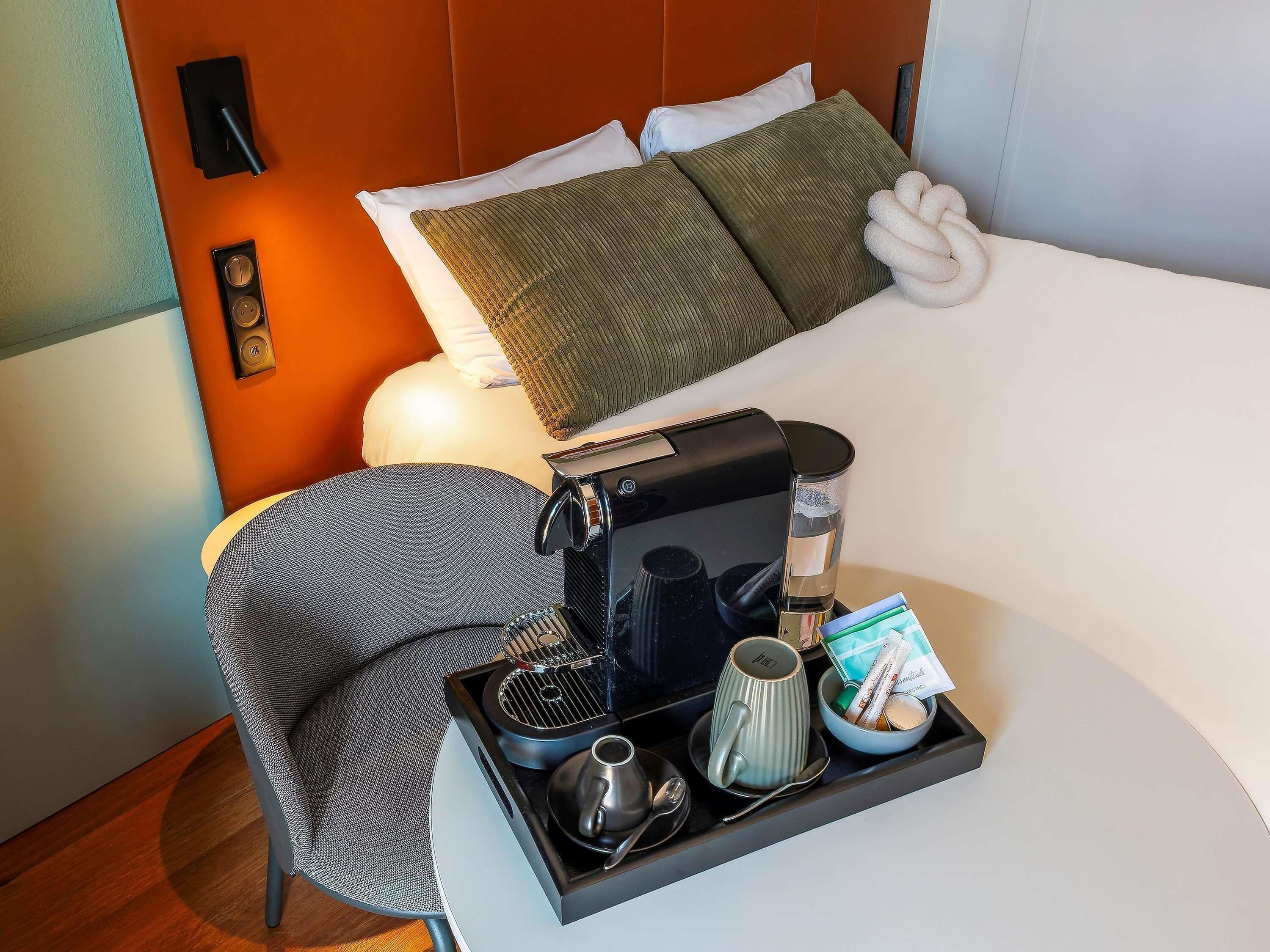 A hotel room with a bed, a bedside table with a coffee maker, a tray with a mug, a bowl of snacks, and a chair.