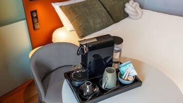 A hotel room with a bed, a bedside table with a coffee maker, a tray with a mug, a bowl of snacks, and a chair.
