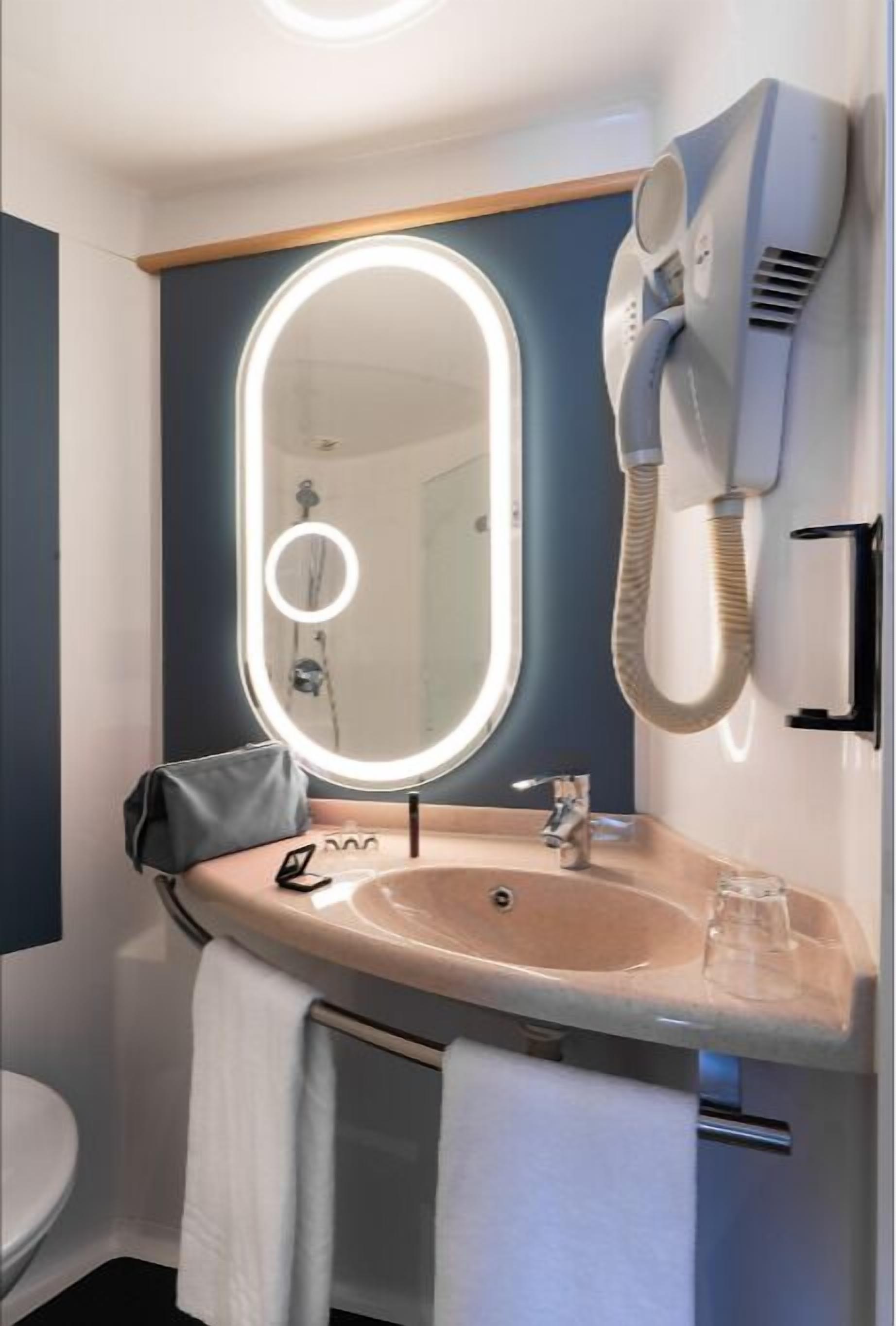 A bathroom with a vanity, a round mirror with lights, a hairdryer, and a towel hanging on the wall.