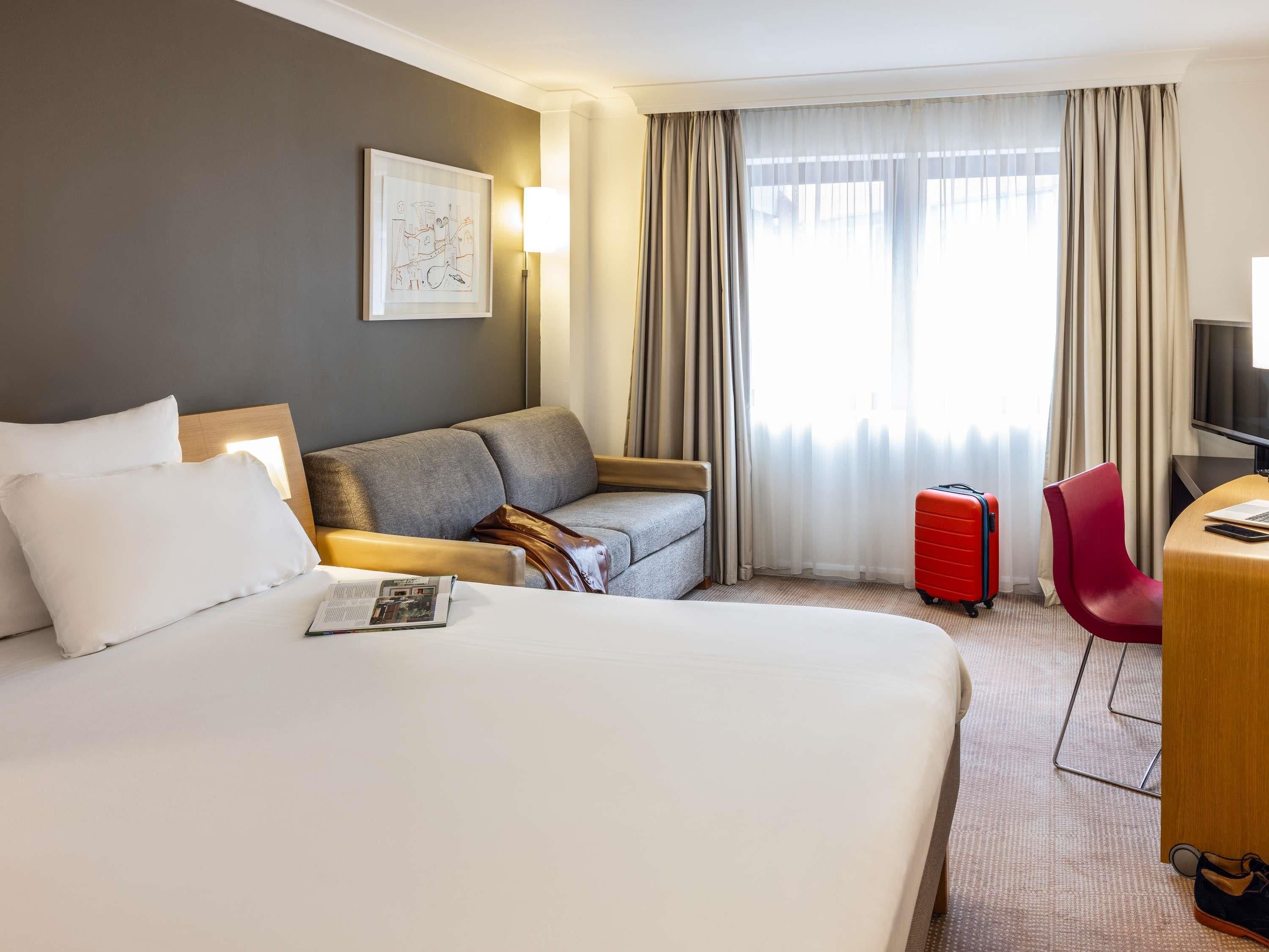 Een hotelkamer met een groot bed, een grijze bank, een rode koffer, een rode stoel en een bureau met een televisie.