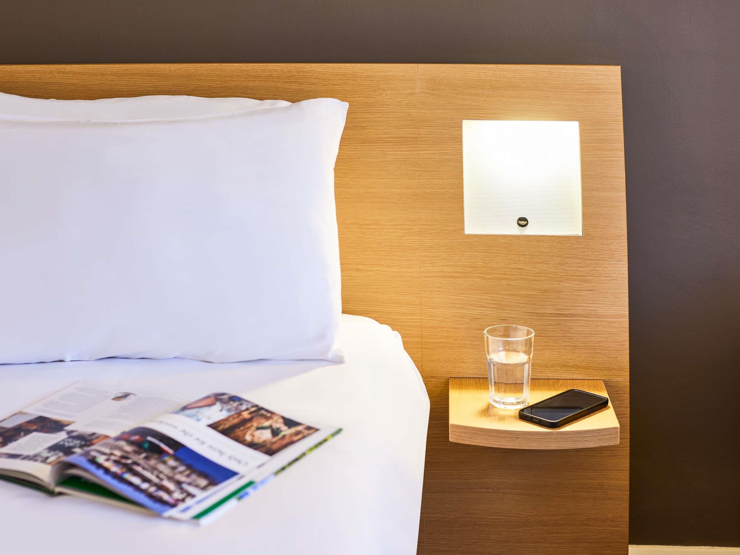 Una cama bien hecha con sábanas blancas, una revista sobre la cama, un vaso de agua y un teléfono inteligente en una mesita de noche de madera.