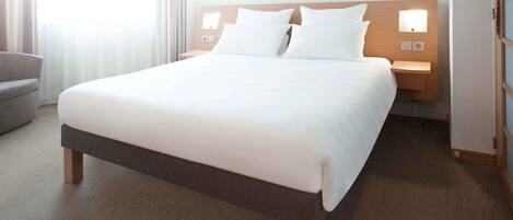 Quarto de hotel com uma cama grande, mesa de cabeceira, cadeira e janela com cortinas.