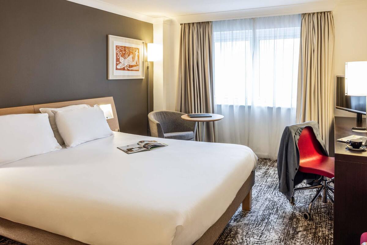 A hotel room with a large bed, a desk, a chair, and a window with curtains.