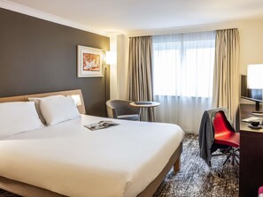 Habitación de hotel con una cama grande, un escritorio, una silla y un ventanal con cortinas.