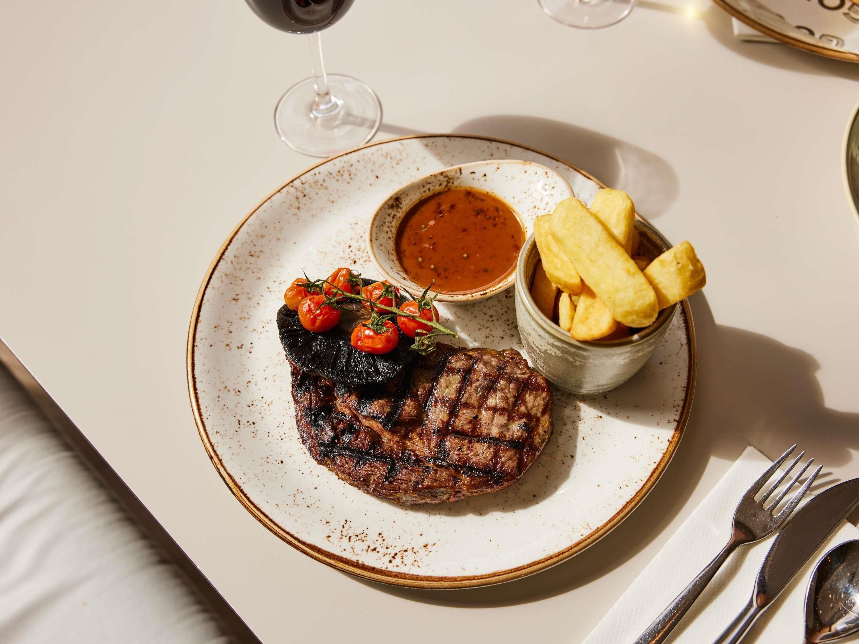 Un plato con bistec a la parrilla, tomates cherry y papas fritas, acompañado de un bol de salsa.