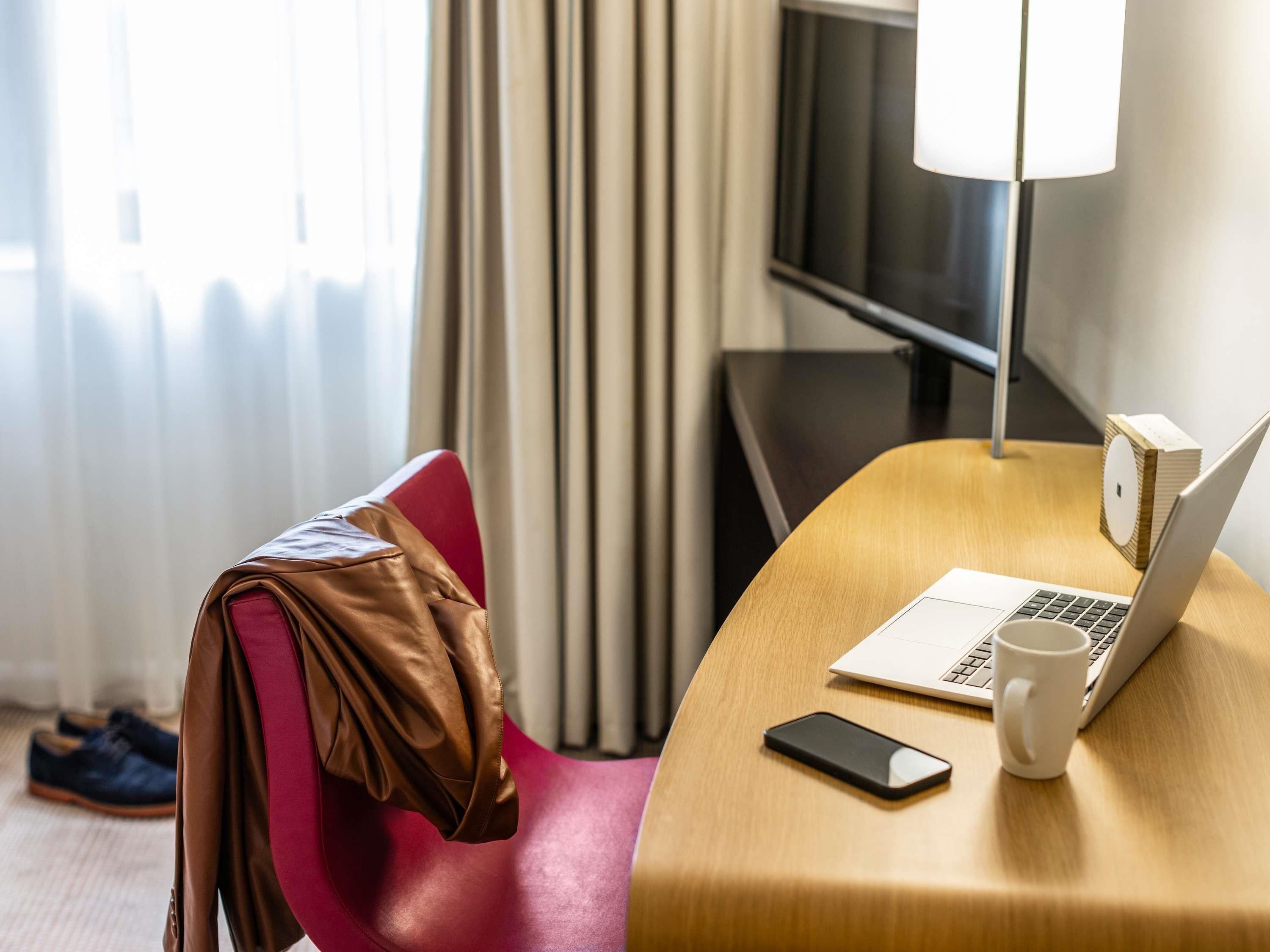 Una habitación de hotel con un escritorio de madera, una computadora portátil, un teléfono inteligente, una taza blanca y una silla roja.