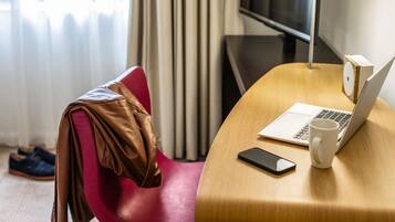 Una habitación de hotel con un escritorio de madera, una computadora portátil, un teléfono inteligente, una taza blanca y una silla roja.
