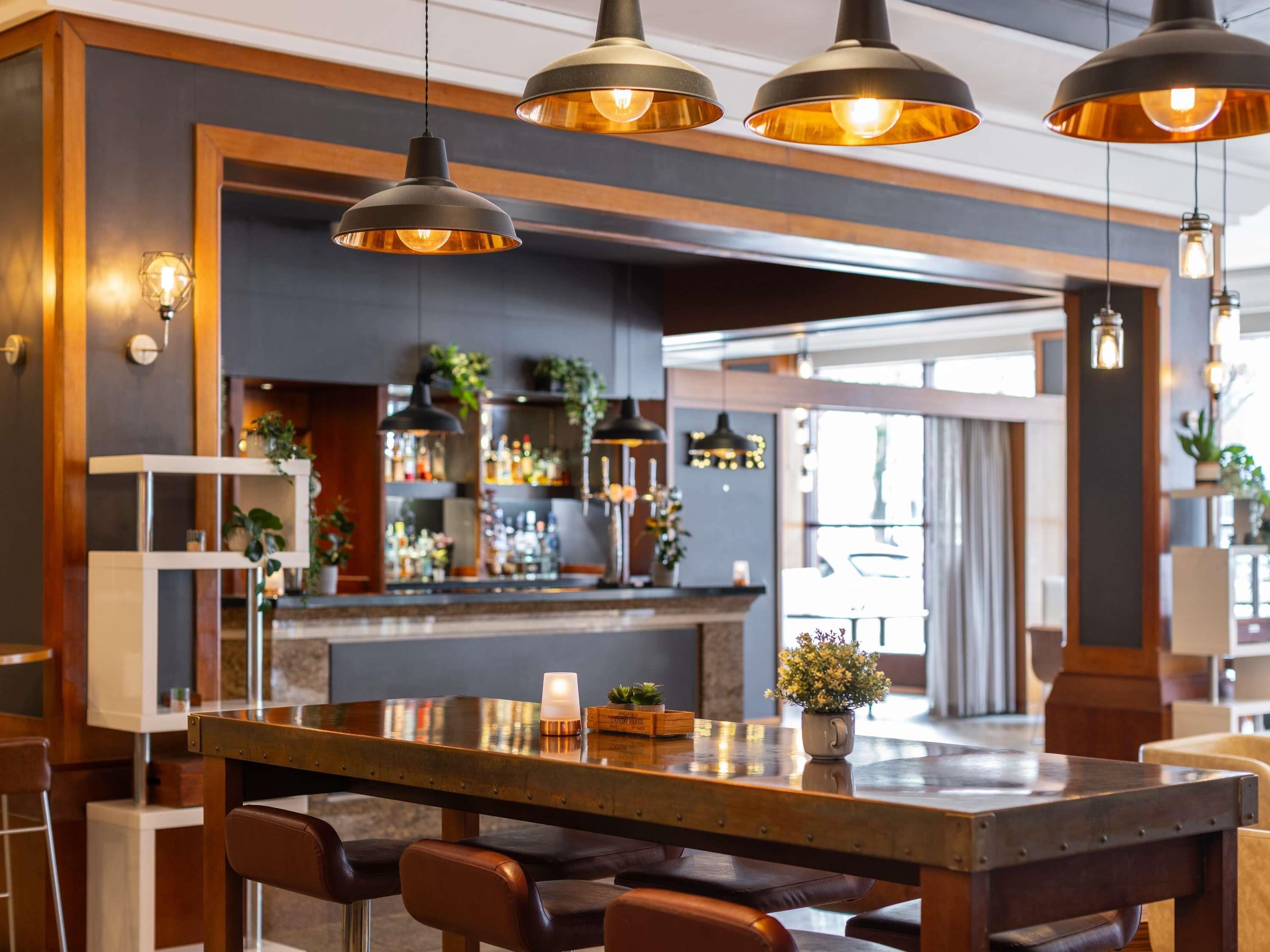 A bar area with a long counter, multiple hanging pendant lights, and a variety of bottles and glasses on shelves.