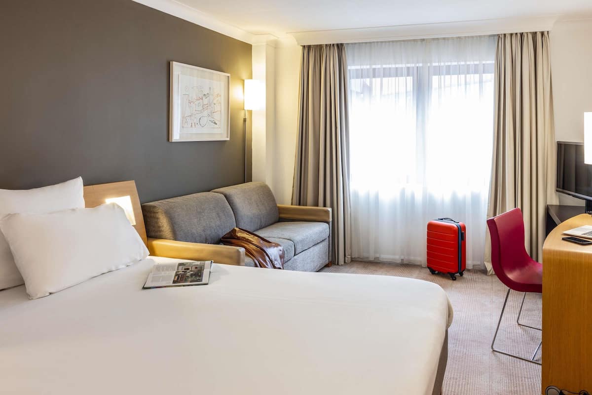 A hotel room with a large bed, a grey sofa, a red suitcase, a red chair, and a desk with a TV.