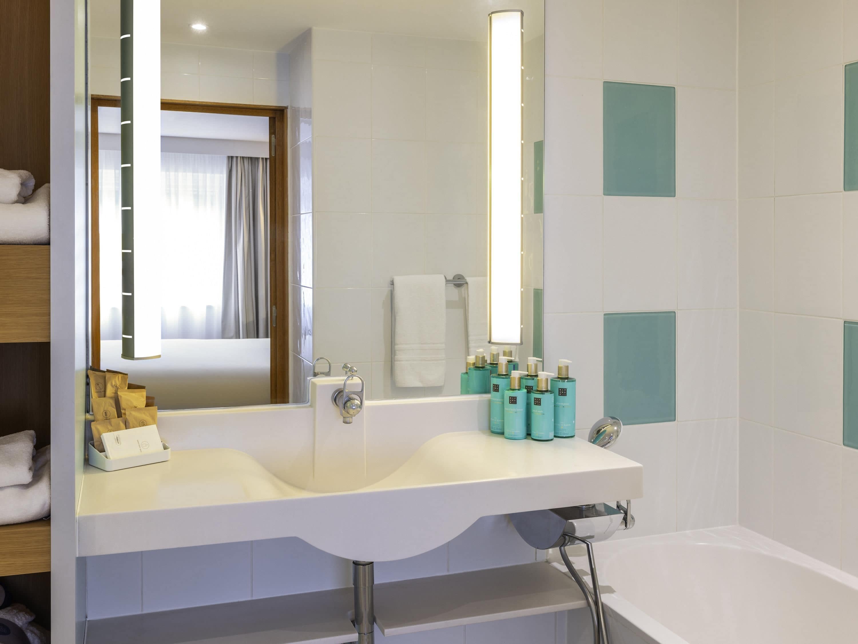 Une salle de bain moderne avec un grand miroir, un lavabo blanc à deux cuves et une baignoire. On trouve des articles de toilette sur le comptoir du lavabo et un porte-serviettes avec des serviettes.