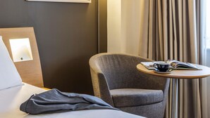 A hotel room with a bed, a gray armchair, a small round table with a book, a cup, and a remote control. - Novotel Bristol Centre (Bristol)