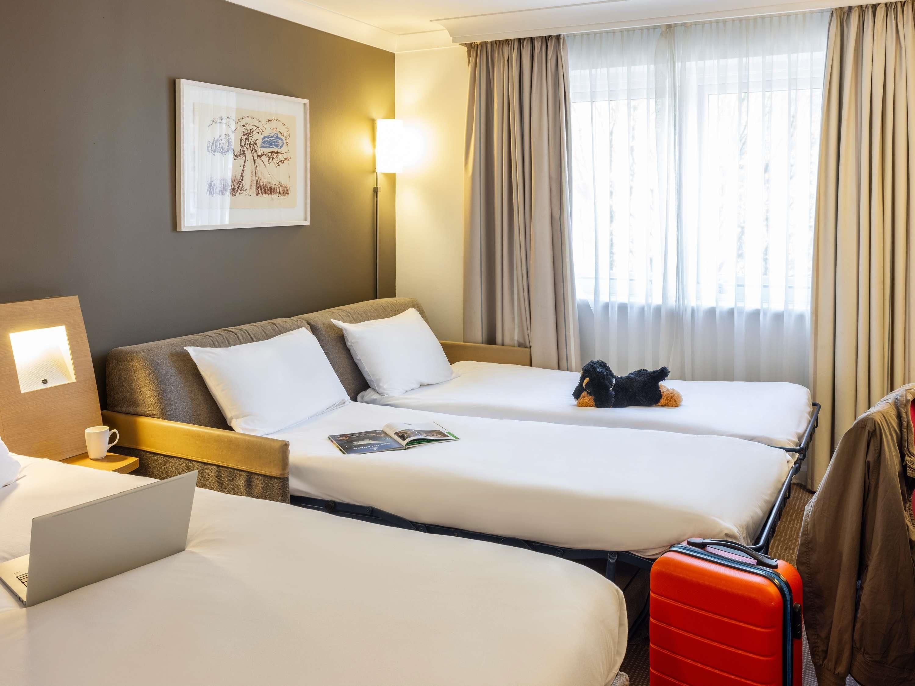 A hotel room with two beds, a desk with a laptop, a framed picture on the wall, and a window with curtains.