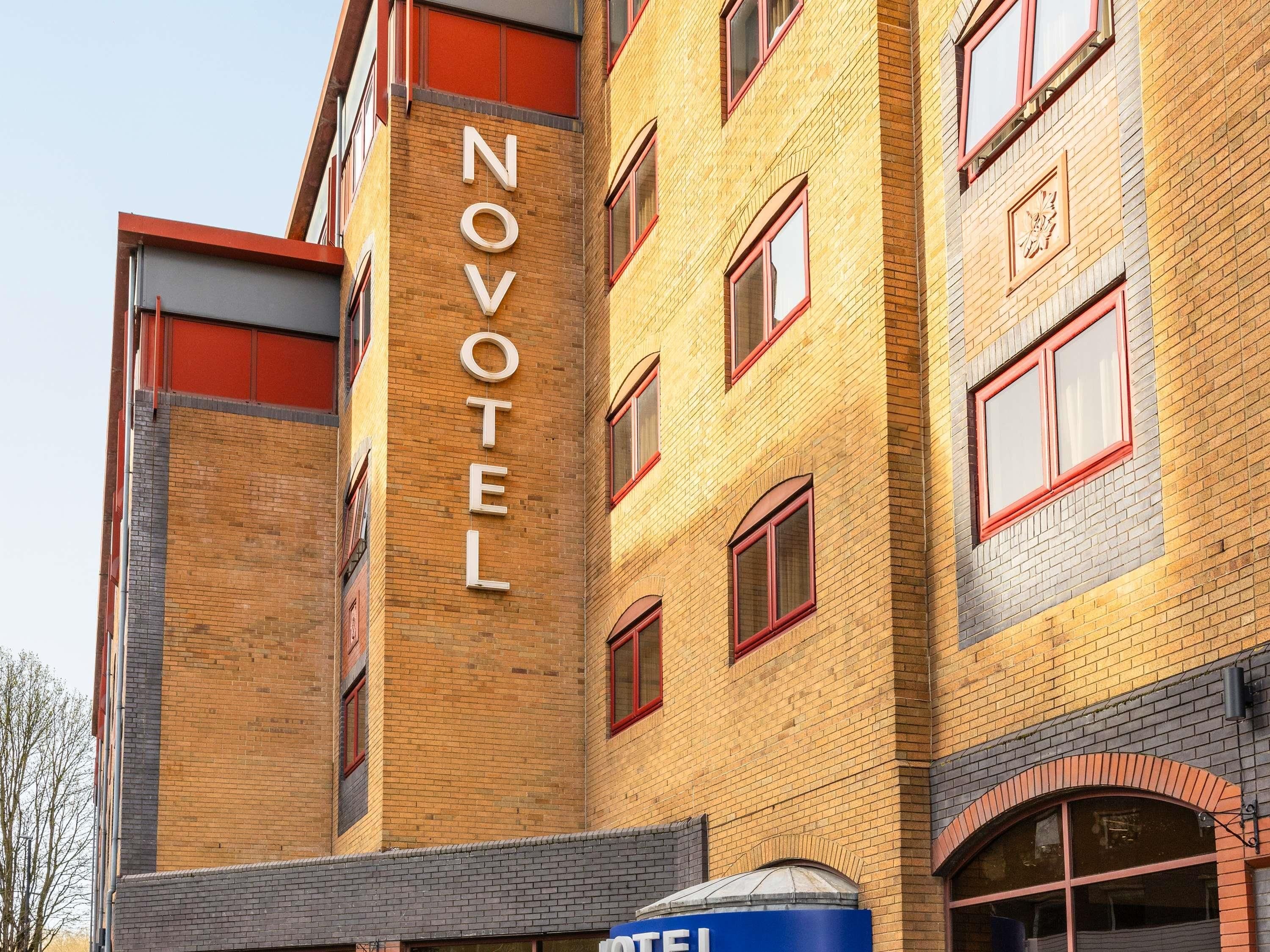 A multi-story hotel with a prominent 'NOVOTEL' sign on the facade, featuring a brick exterior and red-framed windows.