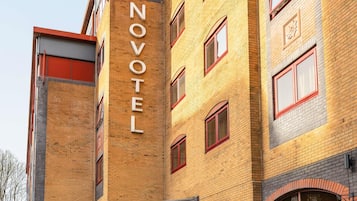 Un hotel de varios pisos con un llamativo letrero 'NOVOTEL' en la fachada, con exterior de ladrillo y ventanas con marco rojo.