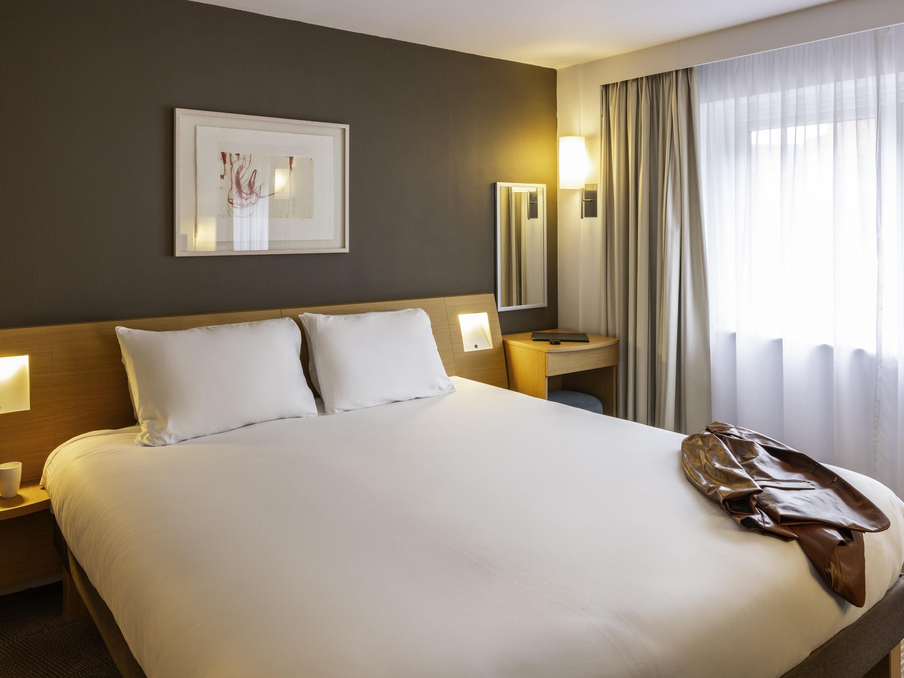 A hotel room with a large bed, bedside tables, a desk, a chair, a window with curtains, and a framed artwork on the wall.