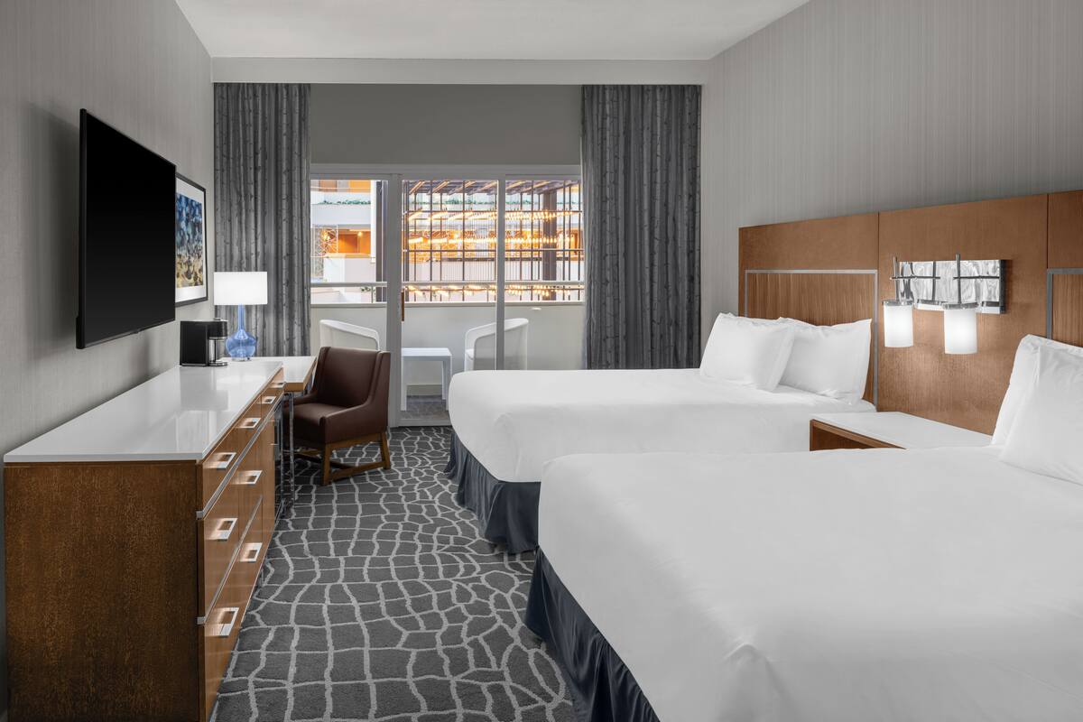 A hotel room with two beds, a desk, a chair, a TV, and a large window with curtains.