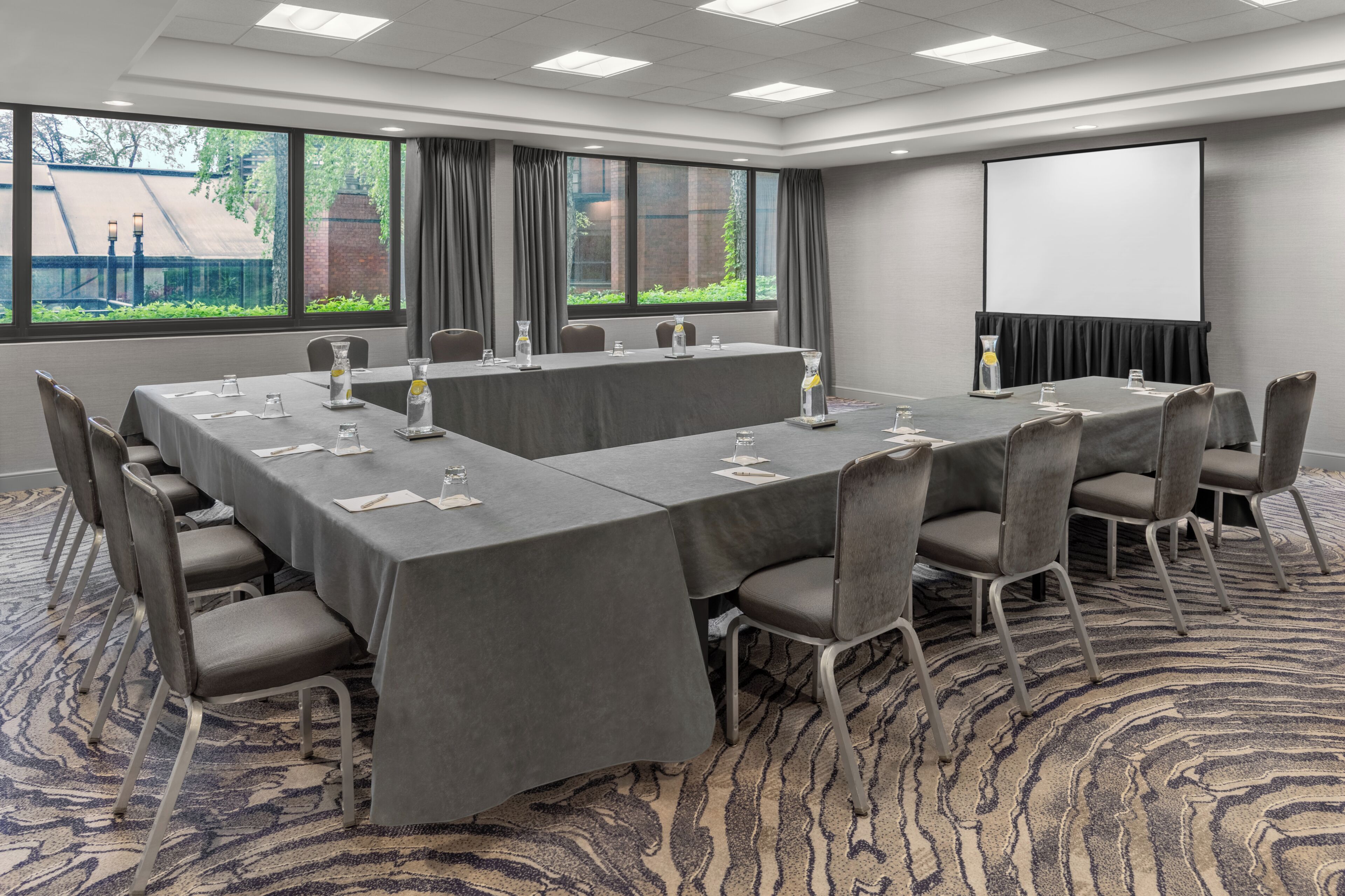 Un salón de conferencias con una larga mesa, sillas, una pantalla de proyector y ventanas amplias.