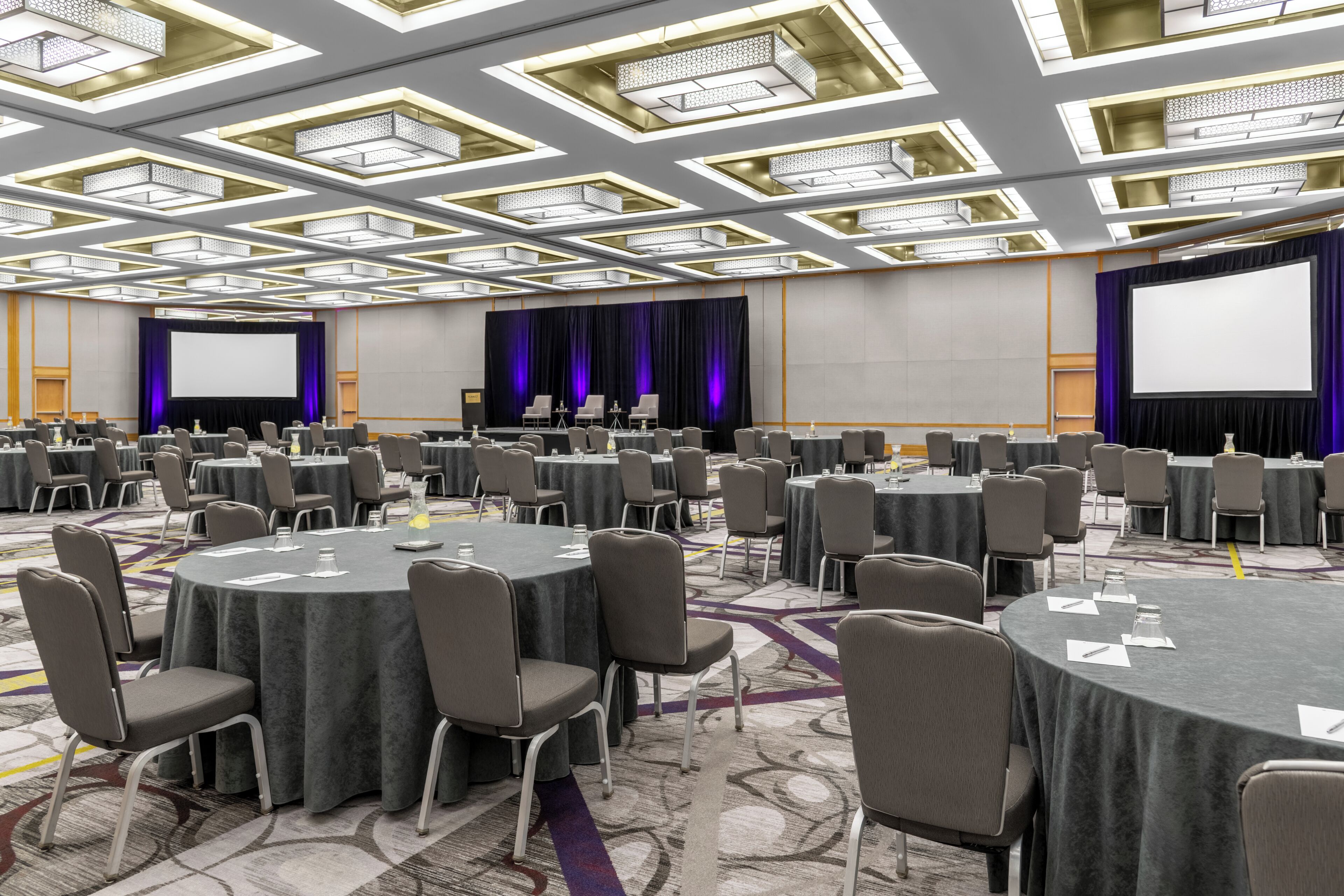 A conference room with round tables, chairs, a stage, and a large screen.