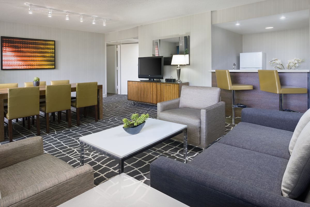 A modern hotel room with a dining area, seating, a flat-screen TV, and a small kitchenette.