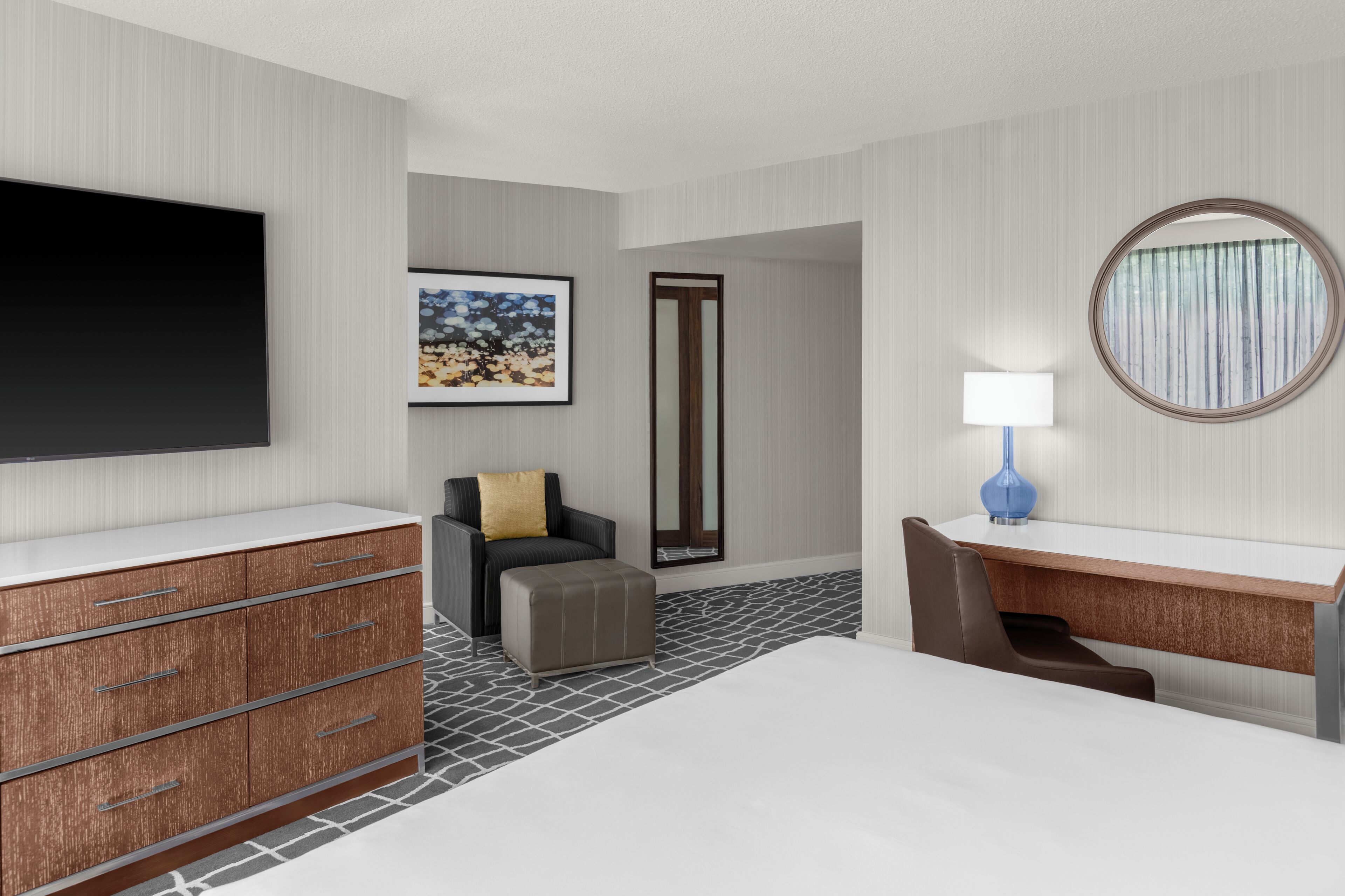 A hotel room with a bed, a desk, a chair, a TV, a framed picture, a mirror, and a lamp.