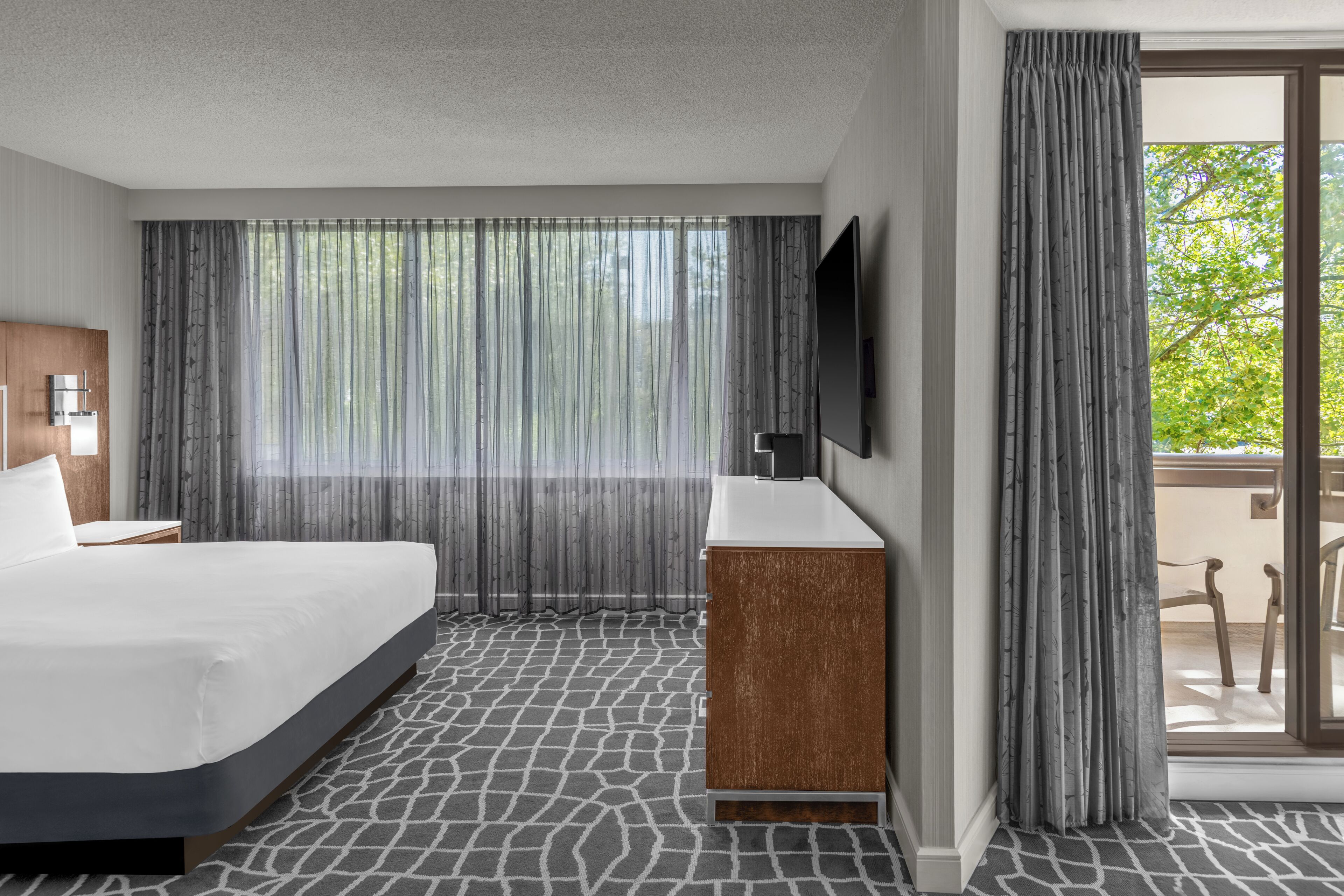 A hotel room with a bed, a wooden cabinet, a flat-screen TV, and a large window with sheer curtains.