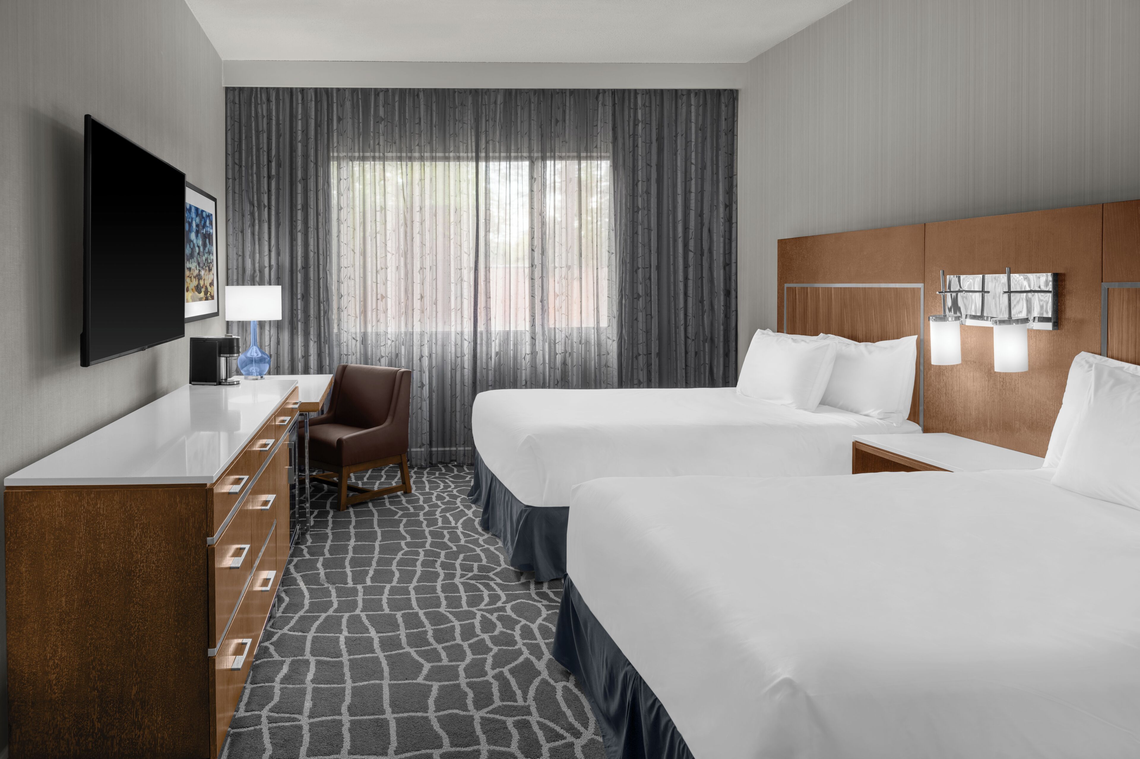 A hotel room with two beds, a TV, a desk with drawers, a chair, a large window with curtains, and a patterned floor.