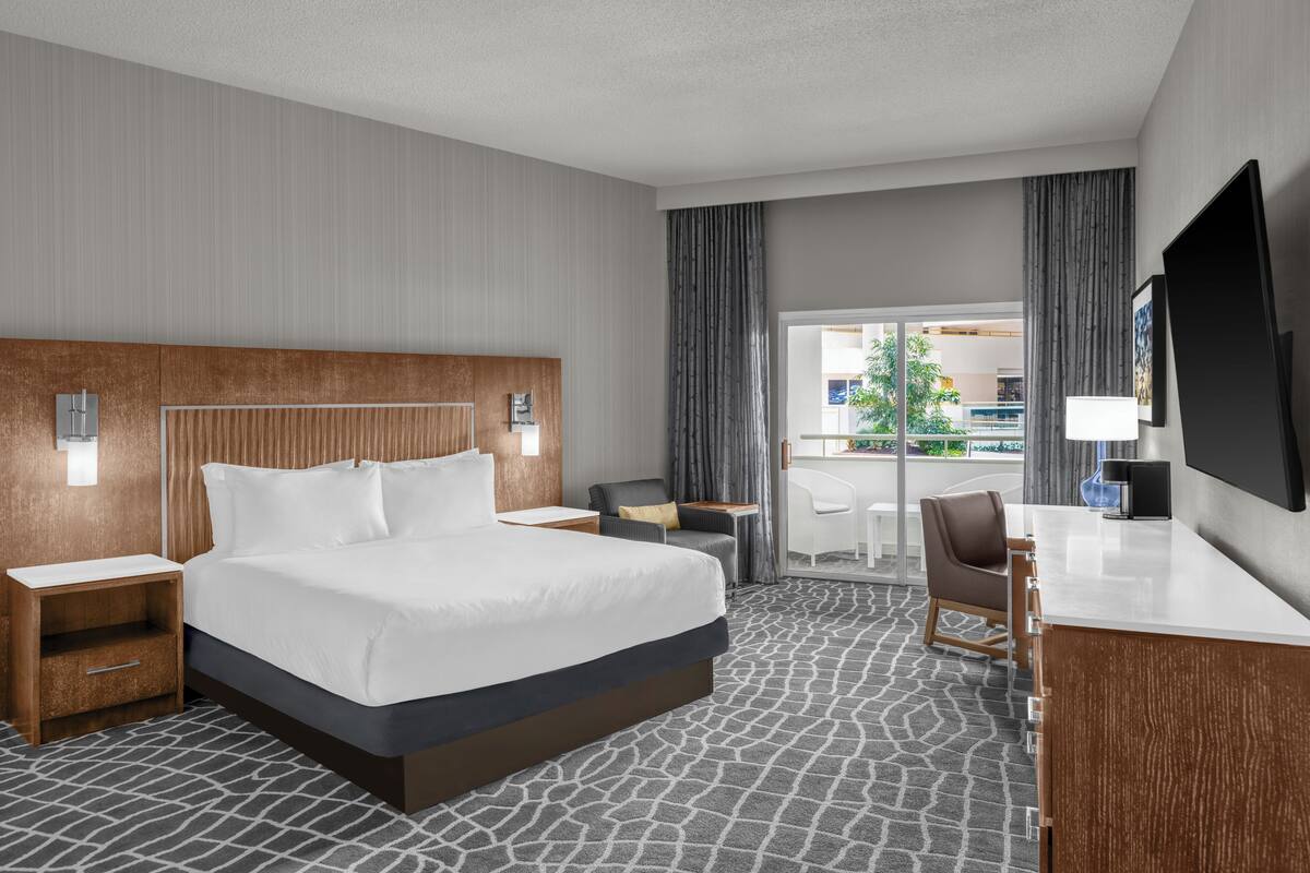 A hotel room with a large bed, a desk, a chair, a TV, and a balcony with a view.