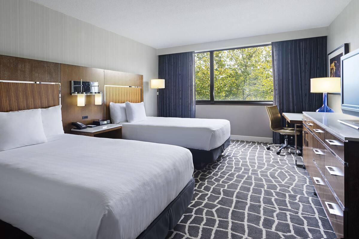A hotel room with two beds, a desk, a chair, a TV, and a window with a view of trees.