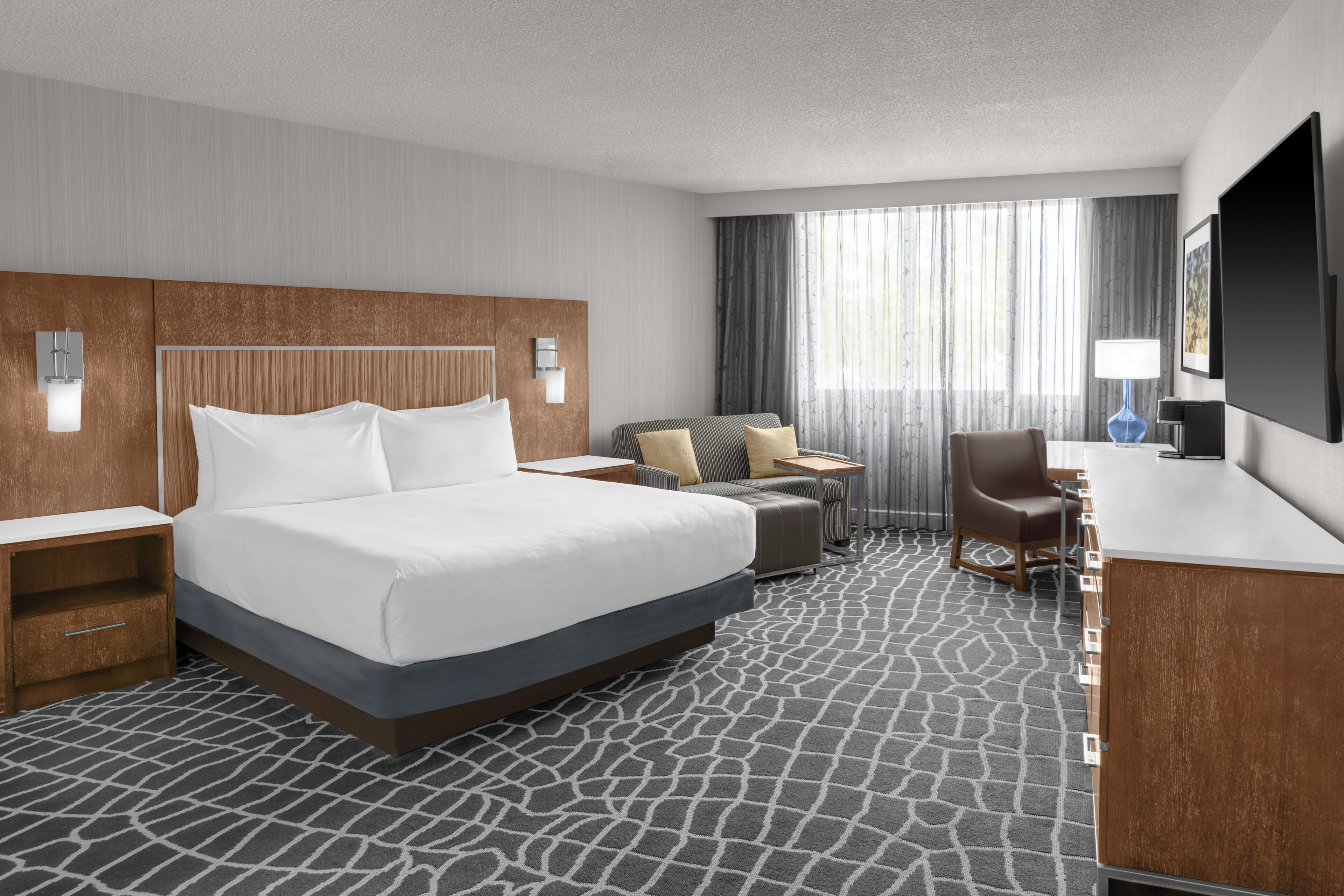 A hotel room with a large bed, a desk, a chair, a TV, and a window with curtains.