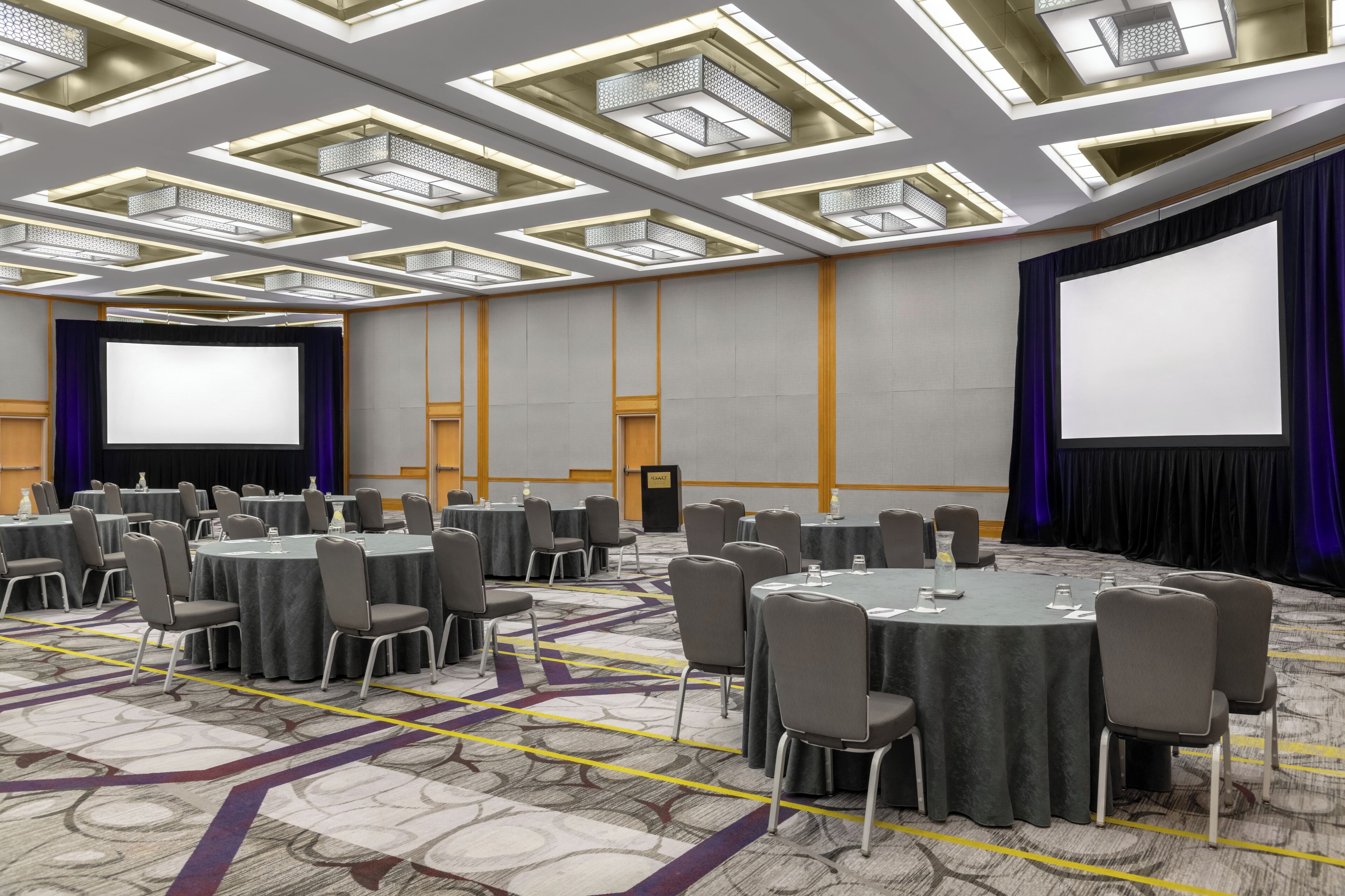 Un salón de conferencias con mesas redondas, sillas, una pantalla de proyector y suelo alfombrado.