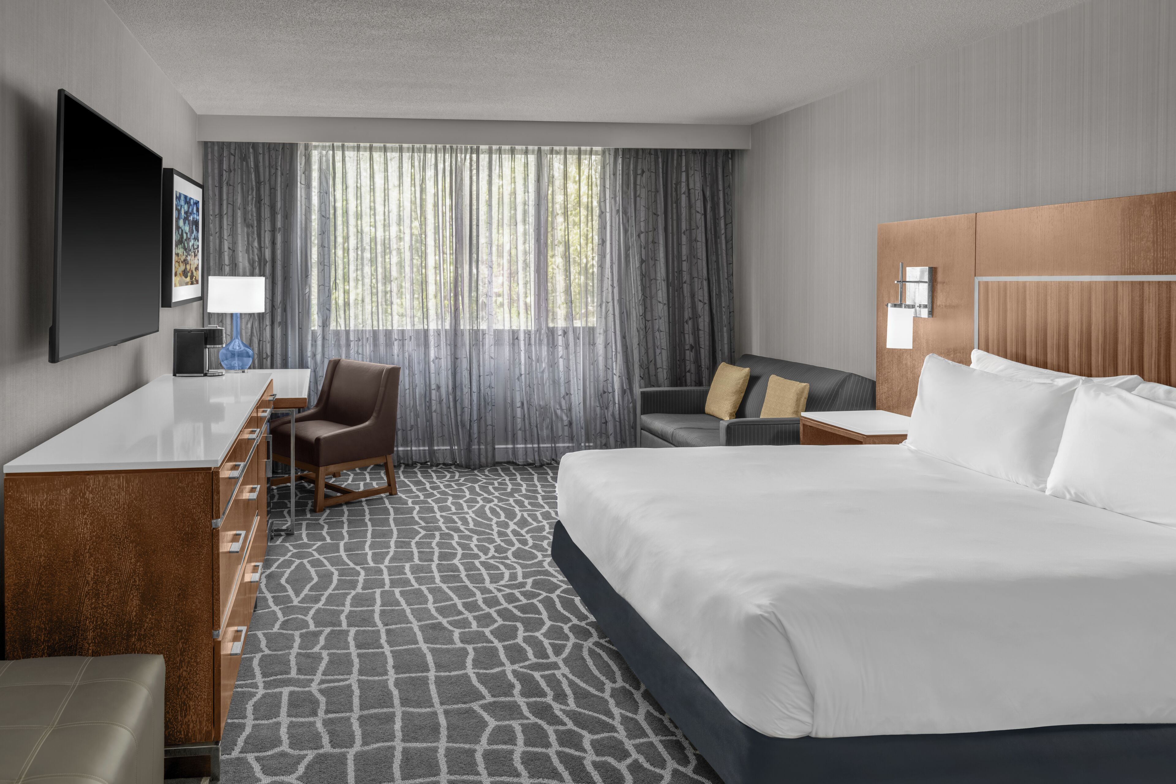 Habitación de hotel con cama, escritorio, silla, televisión y ventana con cortinas.