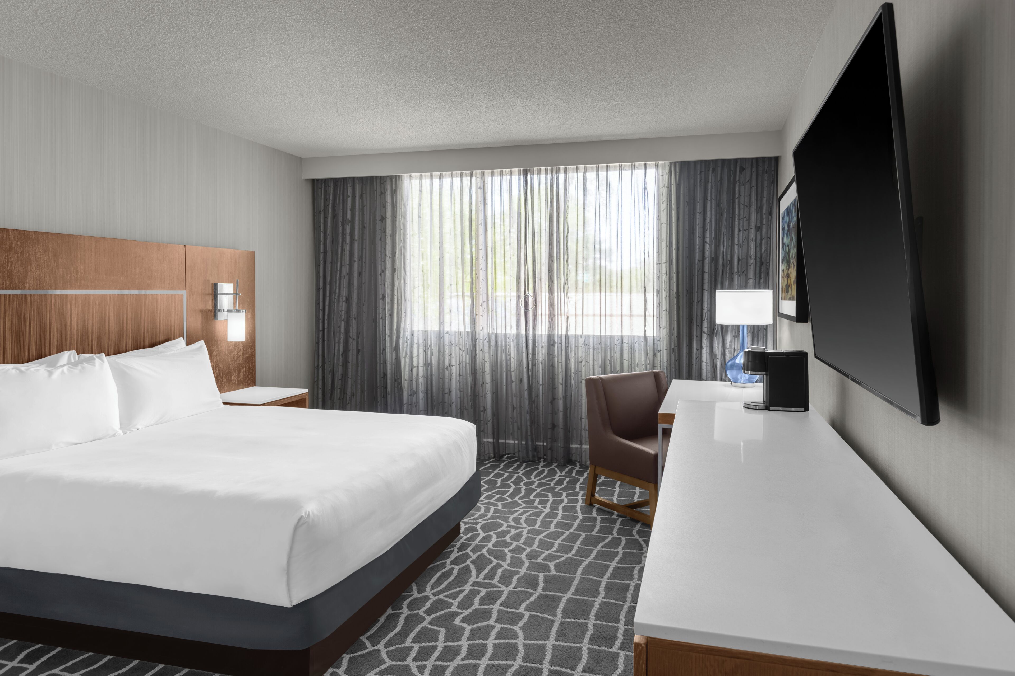 Habitación de hotel con cama, escritorio, silla, televisor y ventana con cortinas.