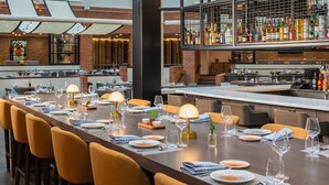 A restaurant with a long dining table set with chairs, a bar area with bottles, and a brick wall in the background. - Hyatt Regency Princeton (Princeton)