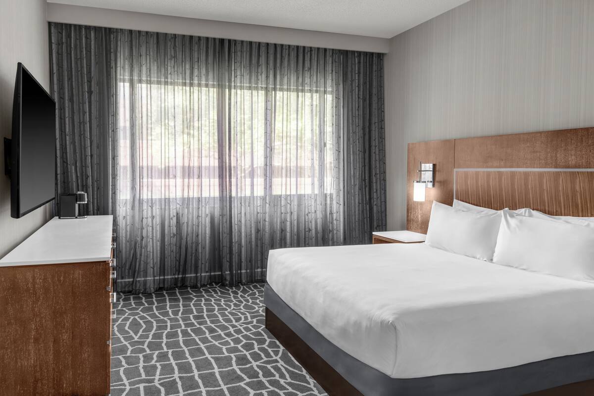 A hotel room with a large bed, a flat-screen TV, a wooden headboard, a nightstand, and a window with sheer curtains.