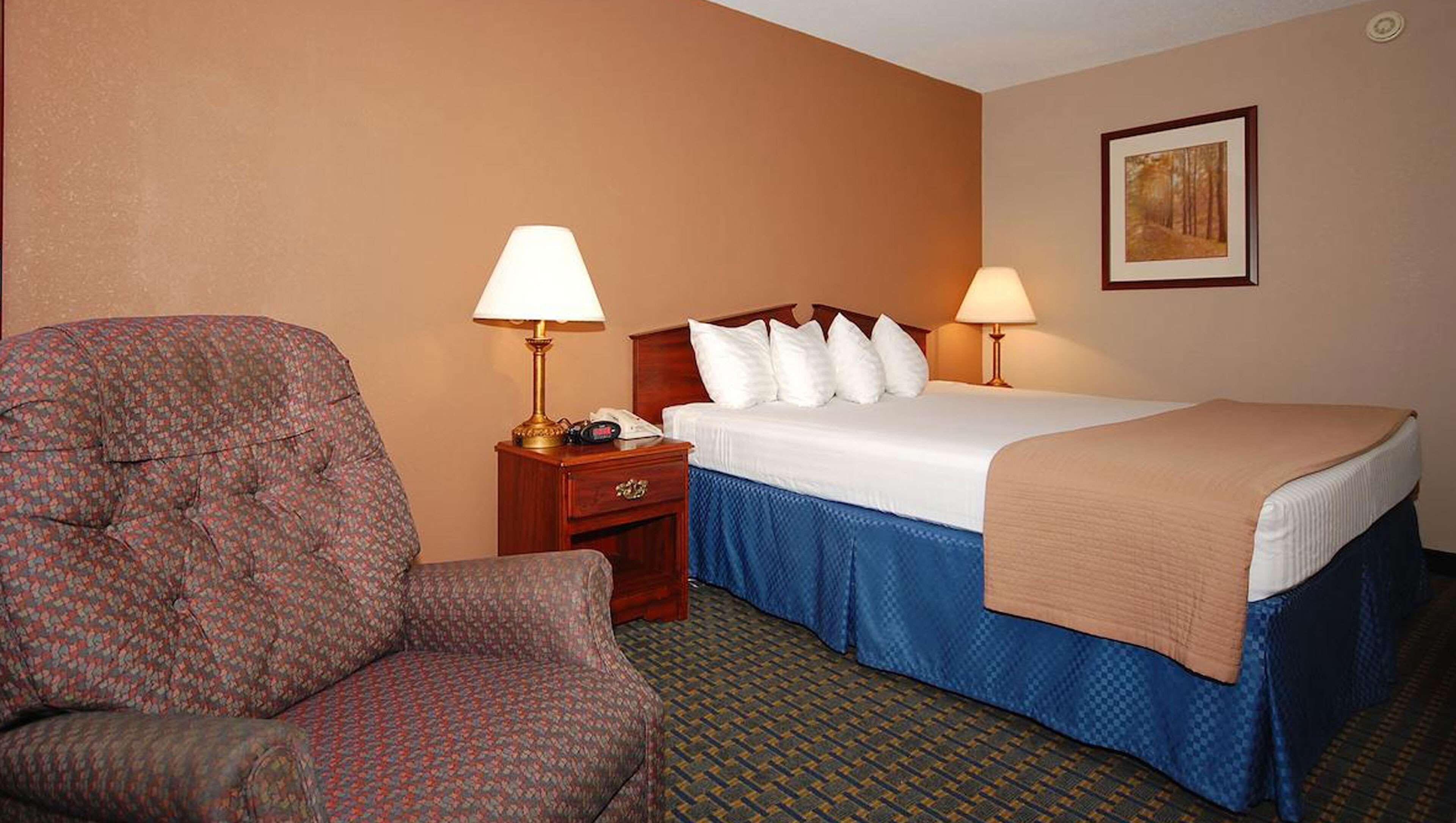A hotel room with a bed, a chair, a nightstand with a lamp, and a painting on the wall.