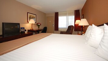 Habitación de hotel con una cama grande, un escritorio con televisor, dos lámparas, una silla y un ventanal con cortinas.